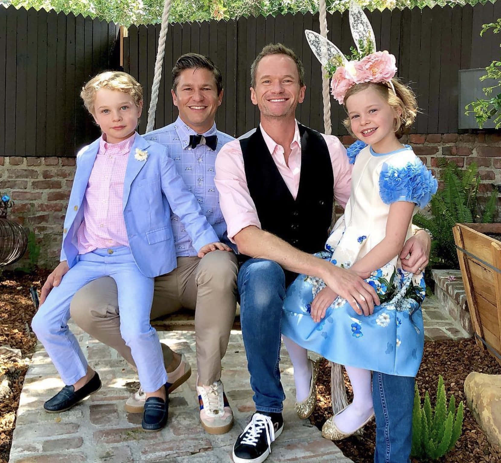 Family 桌布 壁紙 — 四個面帶微笑的人穿著復活節主題服裝，在一個有綠色植物和木柵欄的石頭庭院上擺姿勢。