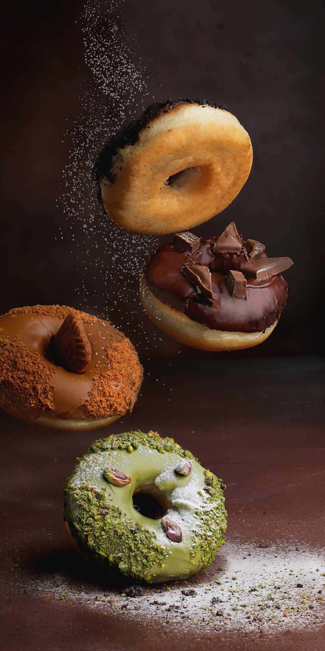 Pastry 桌布 壁紙 — 四個頹廢的甜甜圈懸浮在半空中，糖粉在它們周圍飄落，背景漆黑。
