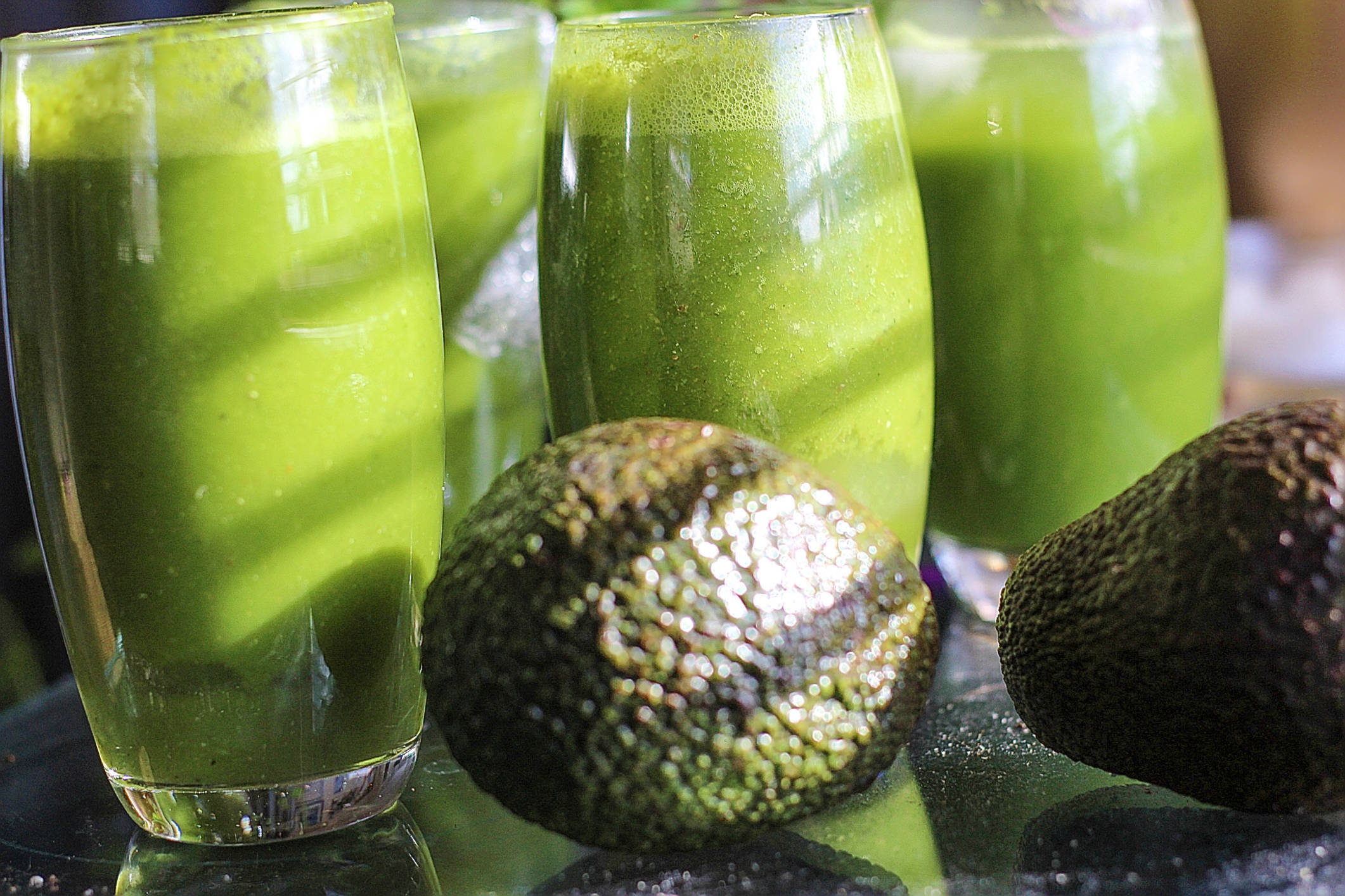 Avocado 桌布 壁紙 — 四個高身玻璃杯裝滿充滿活力的綠色酪梨冰沙，旁邊放著整個酪梨，置於反光表面上。