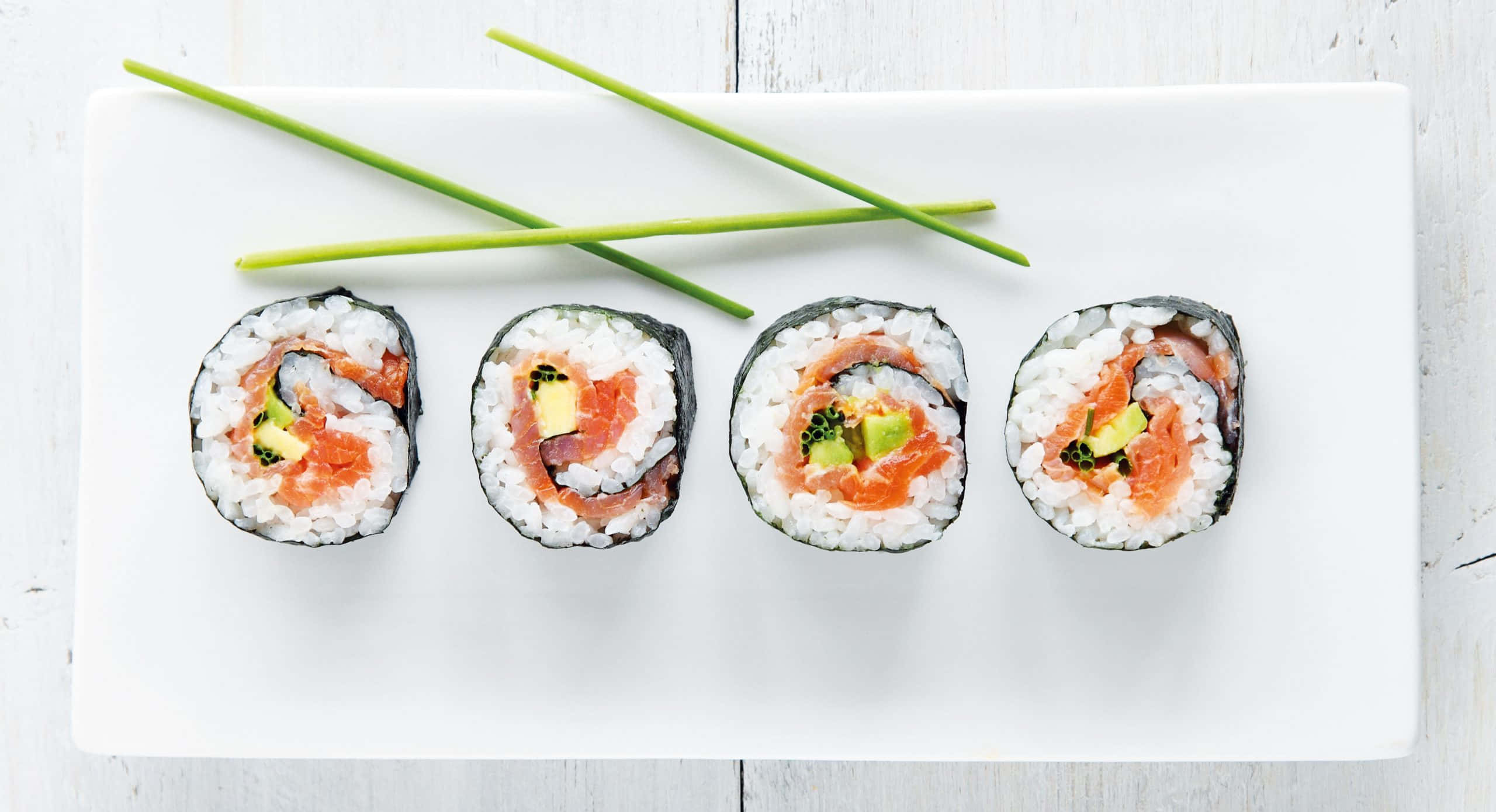 Sushi 桌布 壁紙 — 四個鮭魚酪梨壽司卷整齊排列在白色盤子上，並以交叉的綠色細香蔥點綴。
