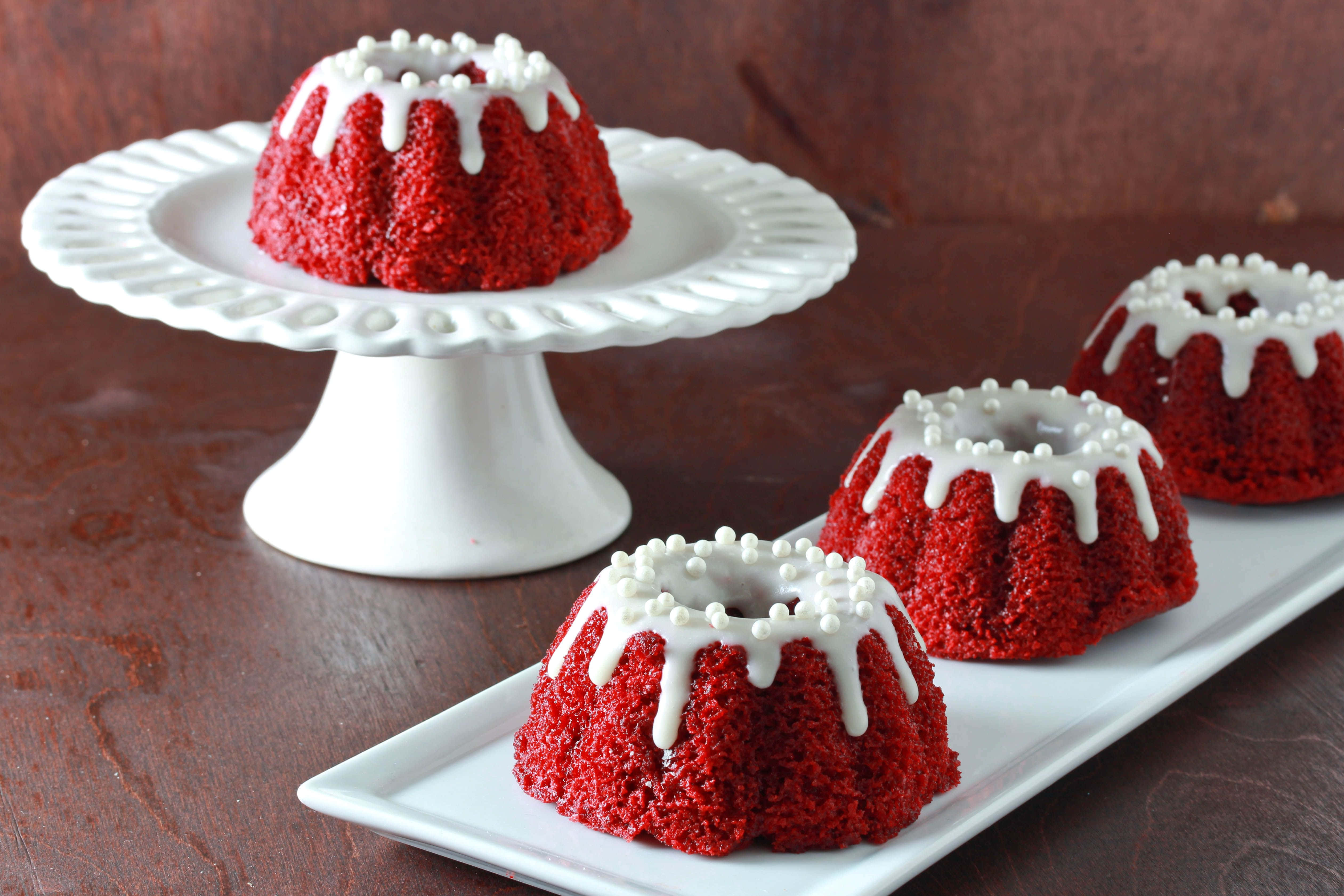 Red Velvet Cake 桌布 壁紙 — 四個鮮紅色的紅絲絨圓環蛋糕，配上白色糖霜和珍珠糖粒，優雅地擺放在白色陶瓷盤上，背景是質樸的木質背景。