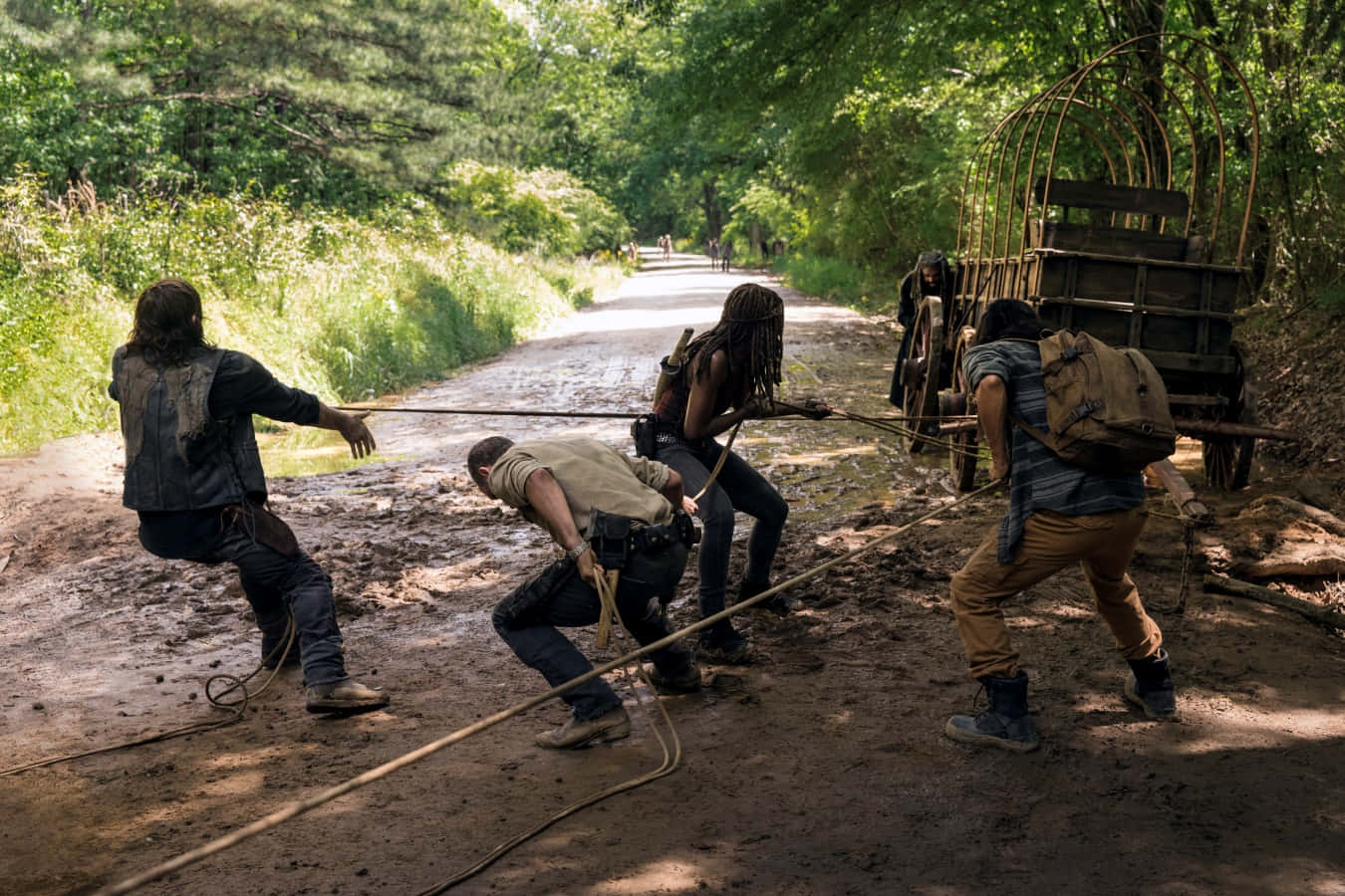 walking dead 桌布 壁紙 — 四名倖存者努力拉著一輛木製手推車，穿過陽光斑駁的森林道路上的厚泥濘。