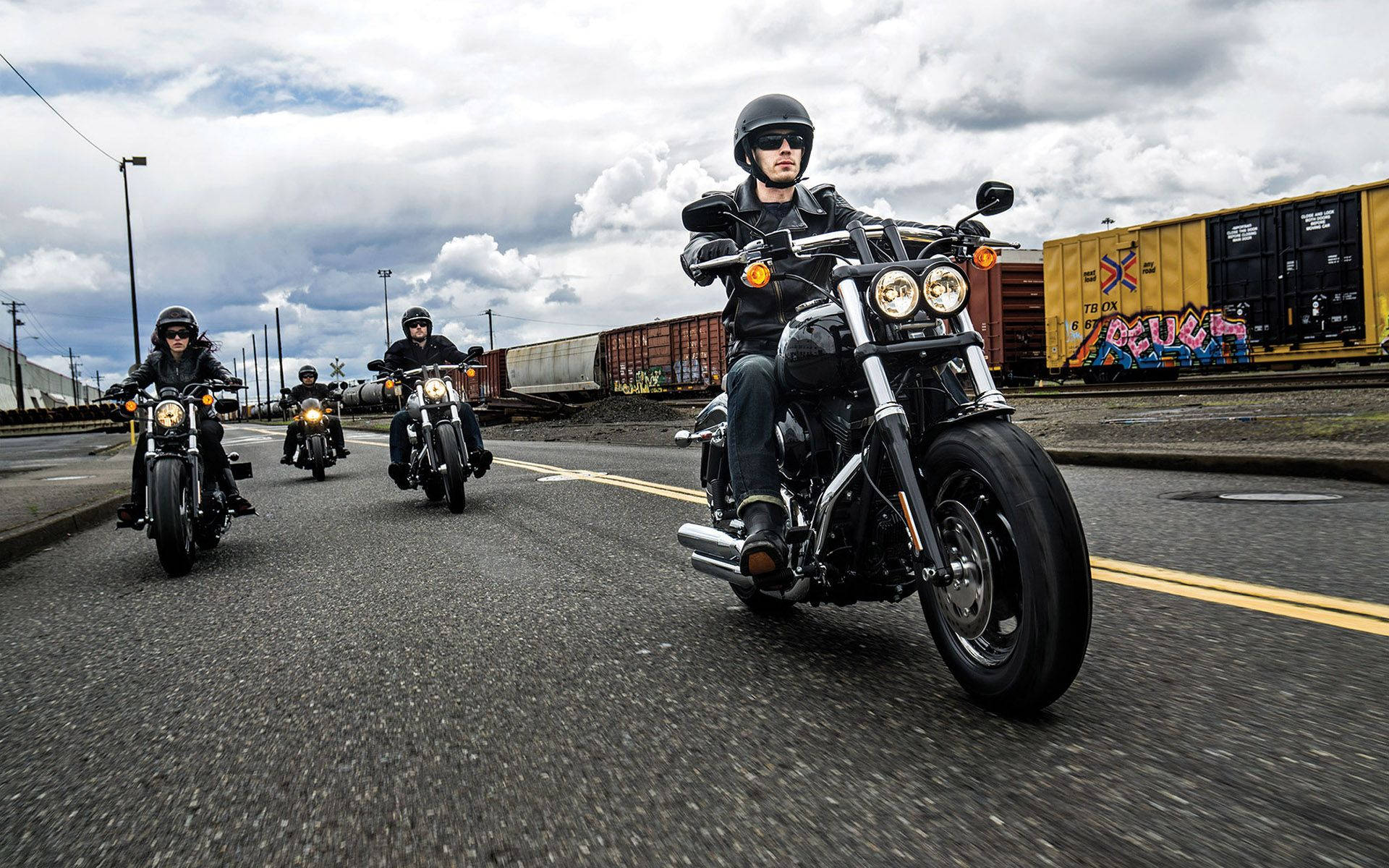 Harley 桌布 壁紙 — 四名摩托車手以隊形在鋪砌的道路上行駛，經過塗鴉覆蓋的貨運火車，天空多雲。