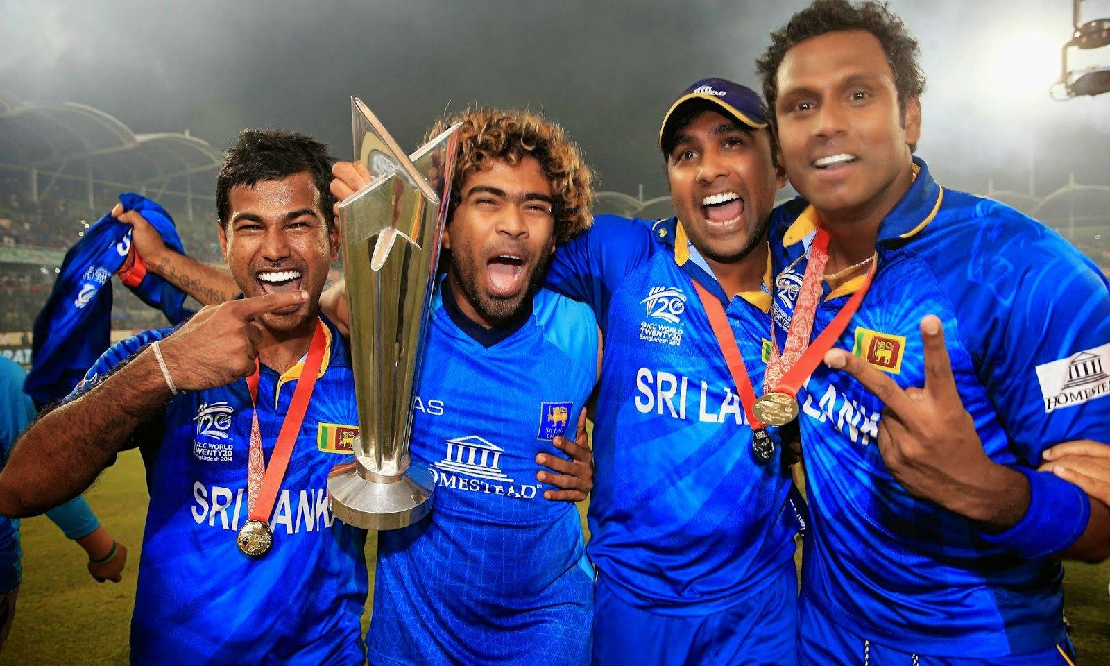 Sri Lanka Cricket 桌布 壁紙 — 四名斯里蘭卡板球運動員身穿藍色球衣，在體育場燈光下高興地舉著銀色獎盃和獎牌慶祝。