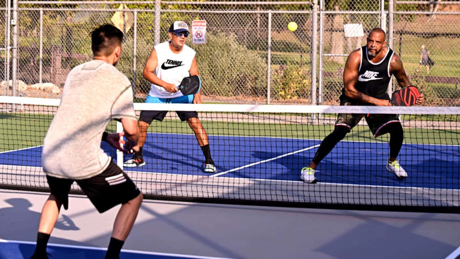 Pickleball 桌布 壁紙 — 四名男子在明亮的藍色球場上激烈地打匹克球，背景有鐵絲網圍欄和綠植。