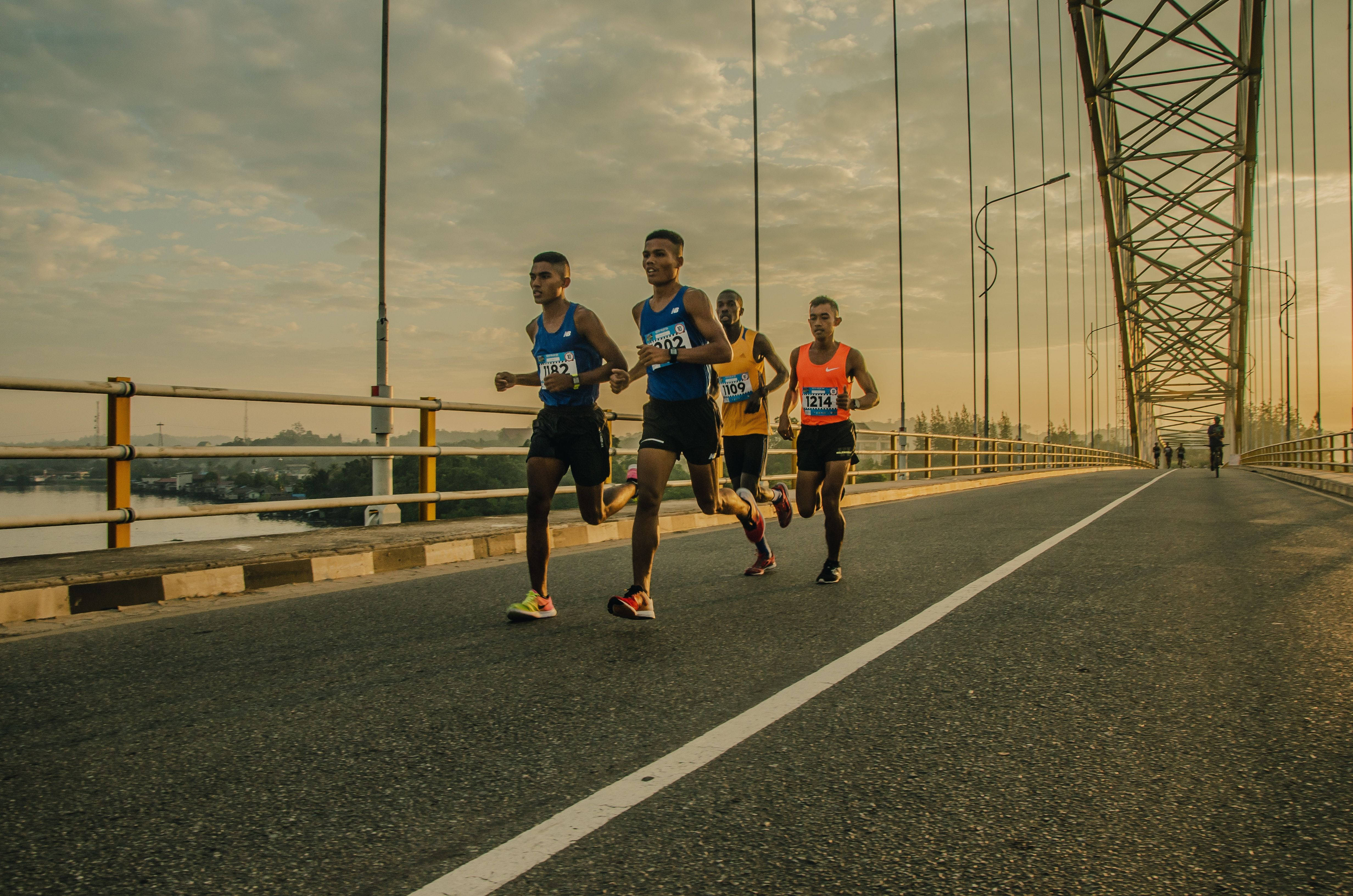 Marathon 桌布 壁紙 — 四名男性跑者穿著運動裝備，在黃金時段的溫暖光線下衝過一條長長的吊橋。
