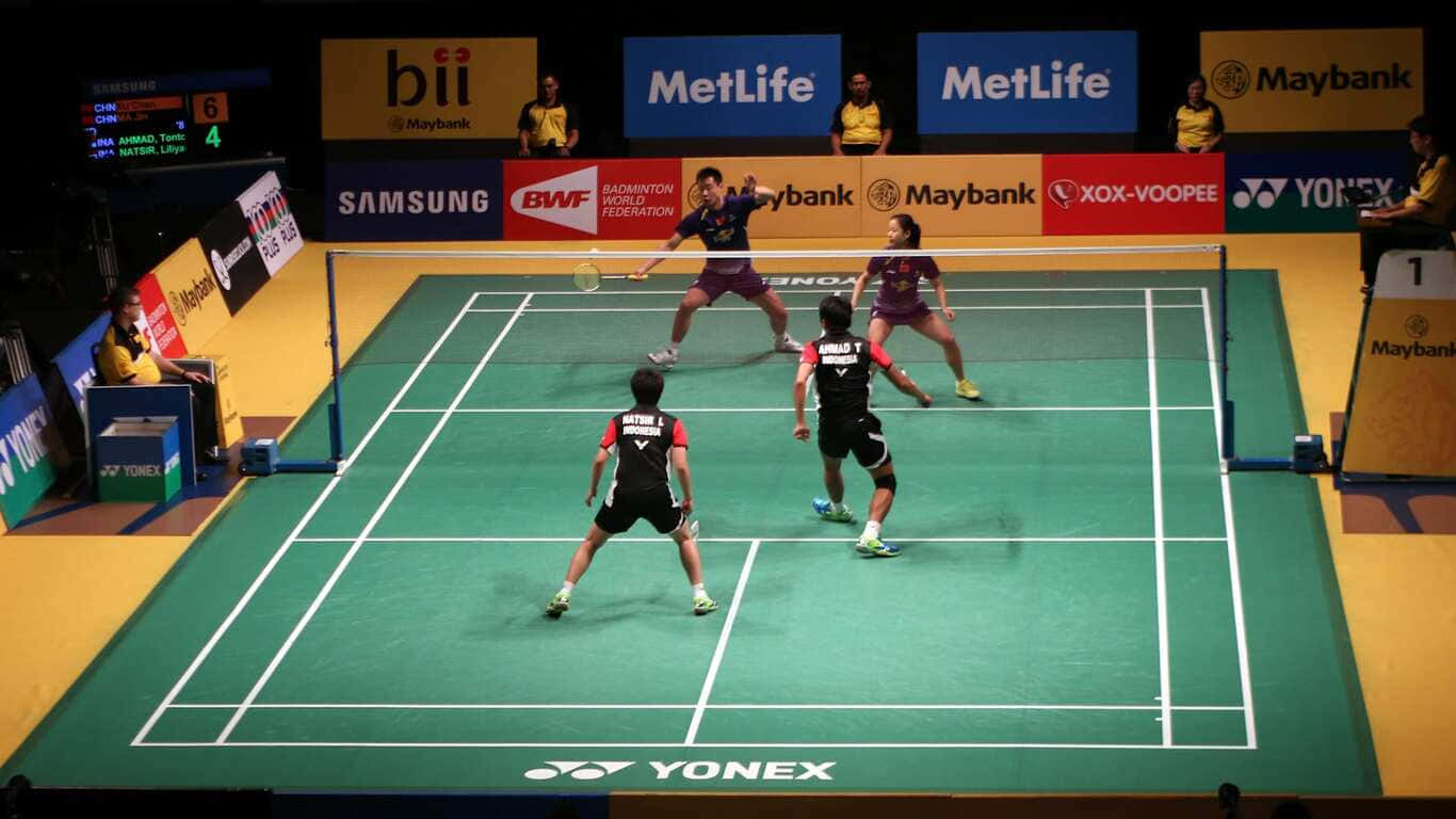 1366x768 Badminton 桌布 壁紙 — 四名羽球運動員在明亮的場館燈光下，於綠色球場上激烈比賽，場邊有品牌橫幅。