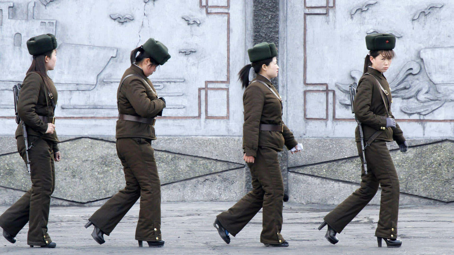 North Korea 桌布 壁紙 — 四名身穿制服的女兵在鋪砌的廣場上列隊行進，背景是帶有雕刻圖案的紋理牆壁。