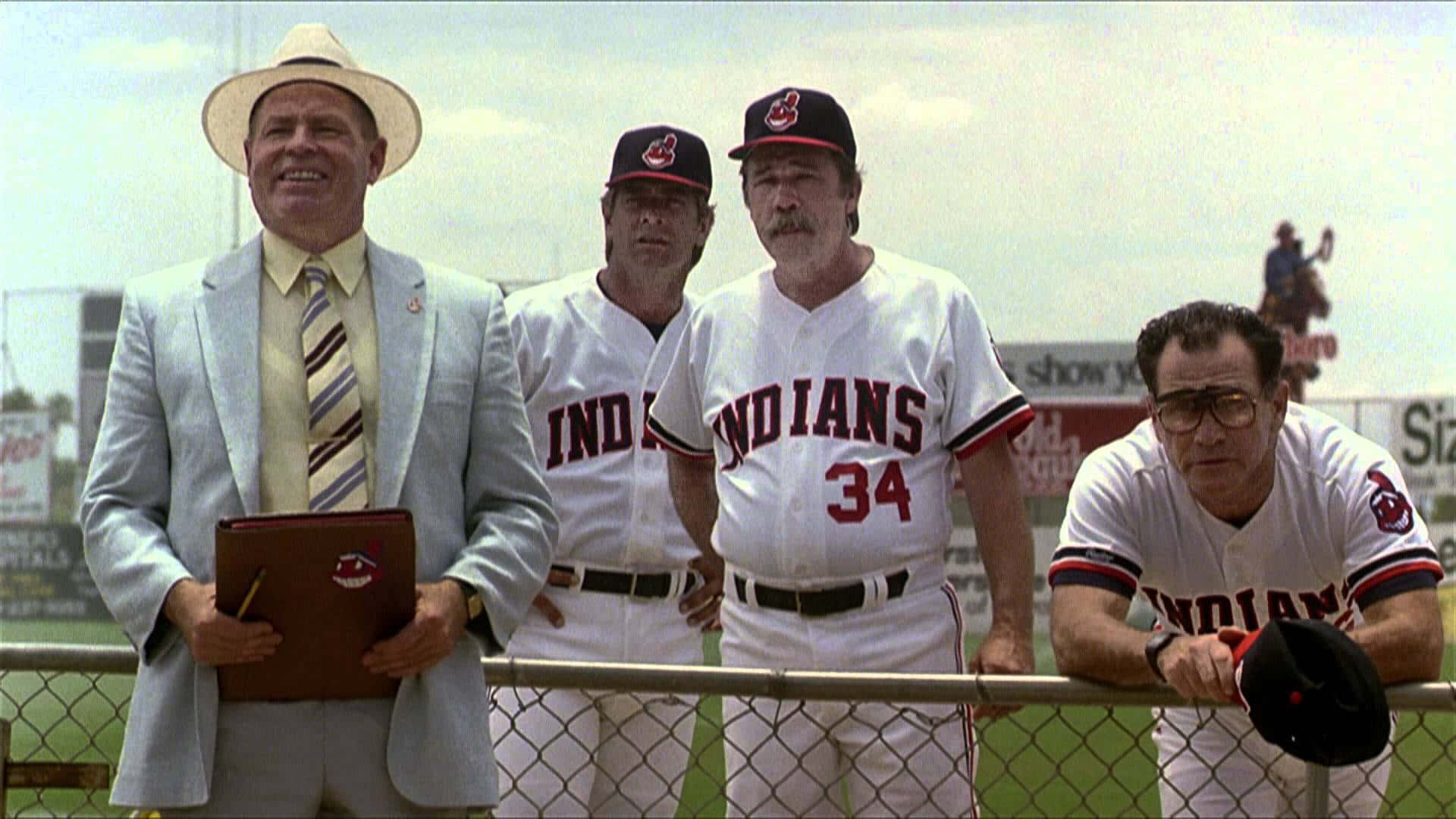 Major League Movie 桌布 壁紙 — 四名身穿棒球制服和西裝的男子站在陽光普照的球場的鐵絲網圍欄旁，遠處可見一名球員。