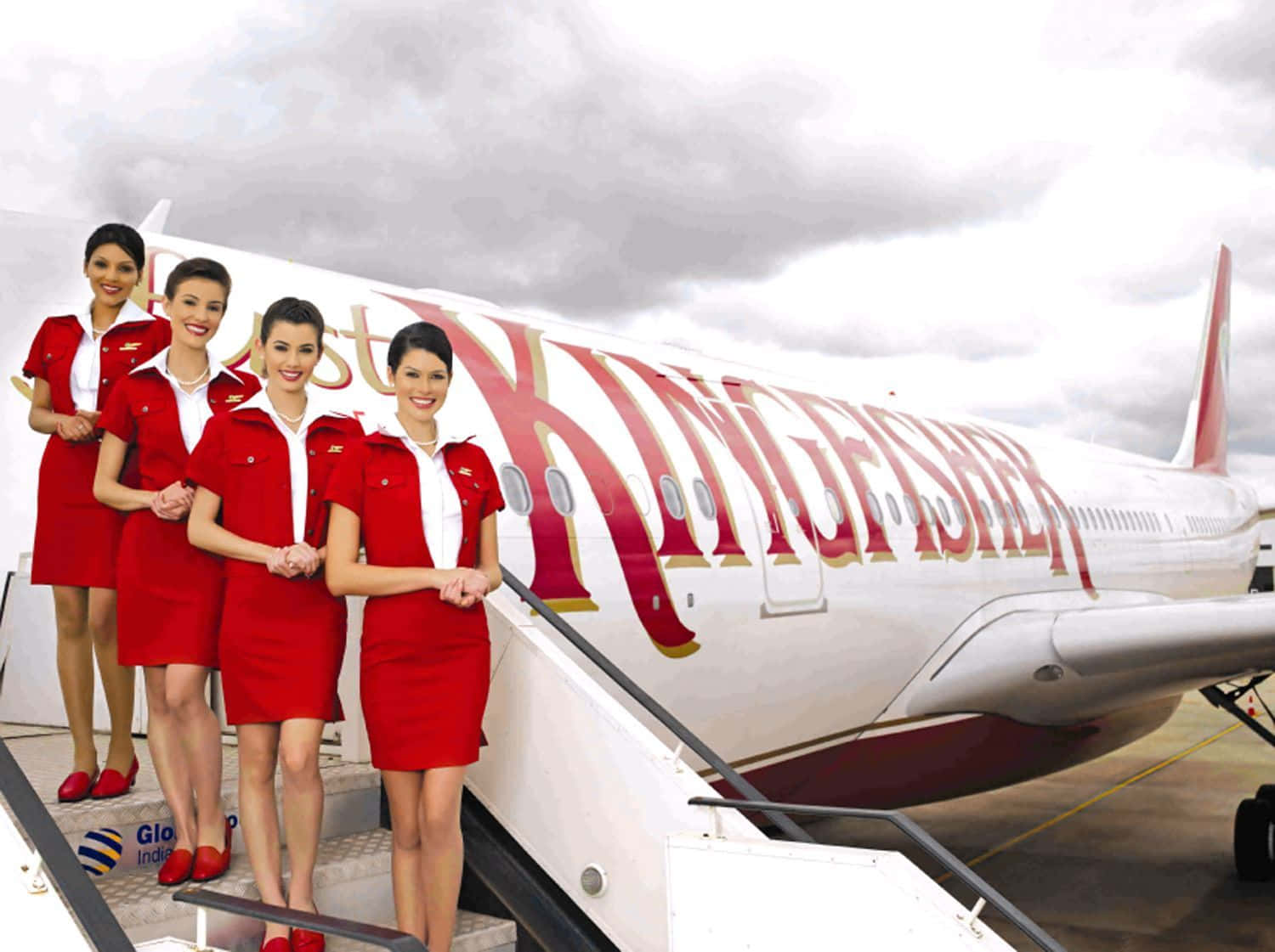 Flight Attendant 桌布 壁紙 — 四名身穿紅色制服、面帶微笑的空服員站在翠鳥航空飛機的樓梯上，天空多雲。