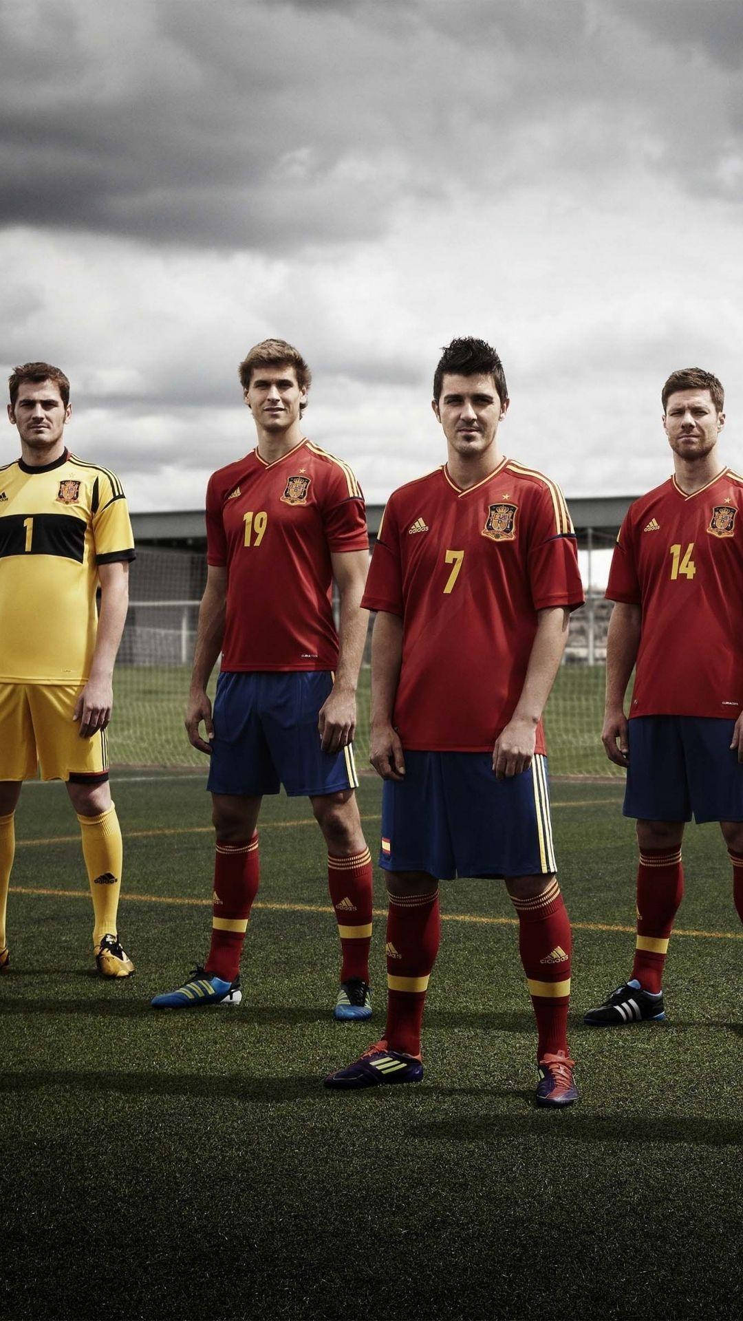 Spain 桌布 壁紙 — 四名身穿紅黃球衣的西班牙足球員自信地站在多雲天空下的綠色草地上。
