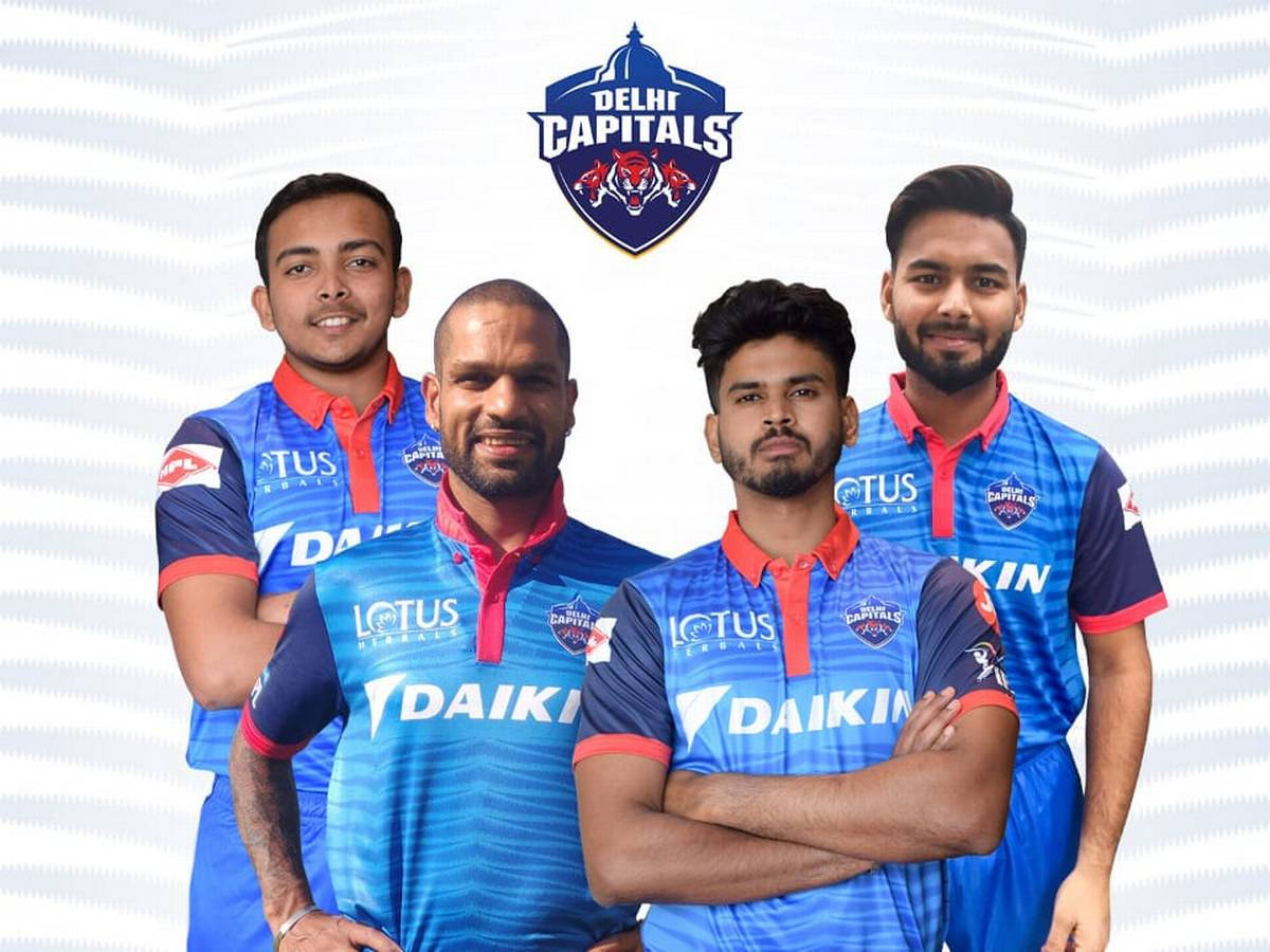 Delhi Capitals 桌布 壁紙 — 四名身穿藍色和紅色德里首都隊球衣的板球運動員在白色波浪形背景前自信地擺姿勢，上方有球隊標誌。