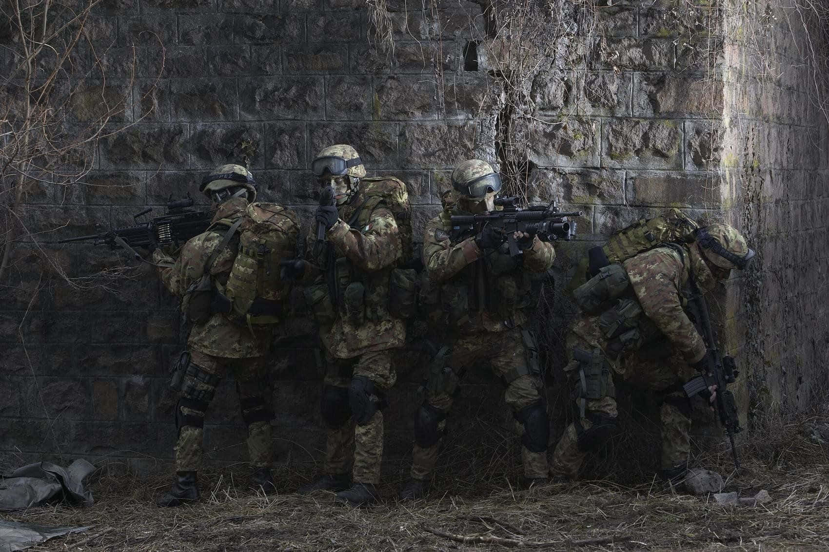  壁紙 — 四名身穿迷彩裝備的士兵蹲在風化的石牆後面，手持步槍，處於緊張的戰鬥姿態。