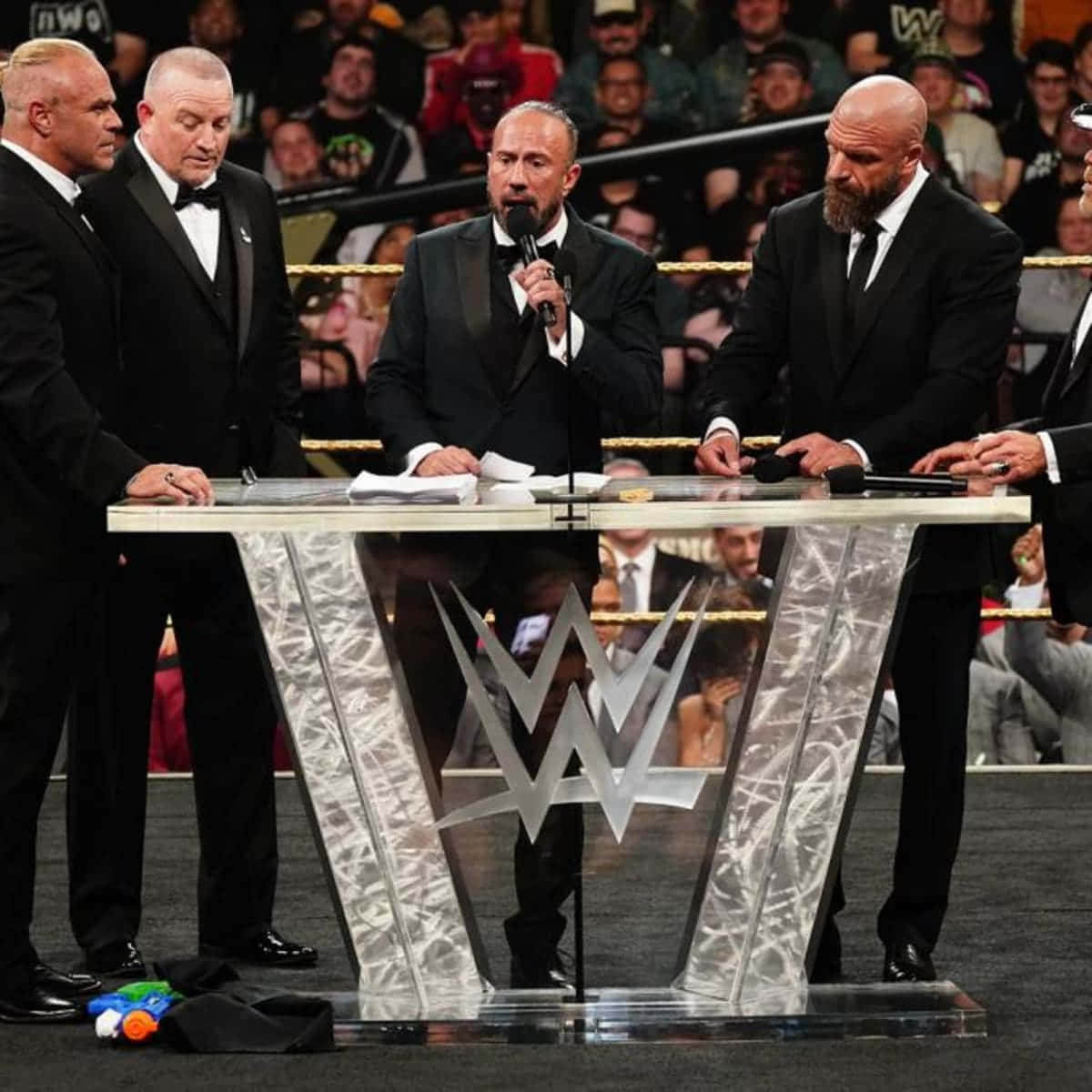 triple h 桌布 壁紙 — 四名身穿黑色燕尾服的男子莊嚴地站在一個帶有標誌的透明WWE講台周圍，向摔角場館中的群眾講話。