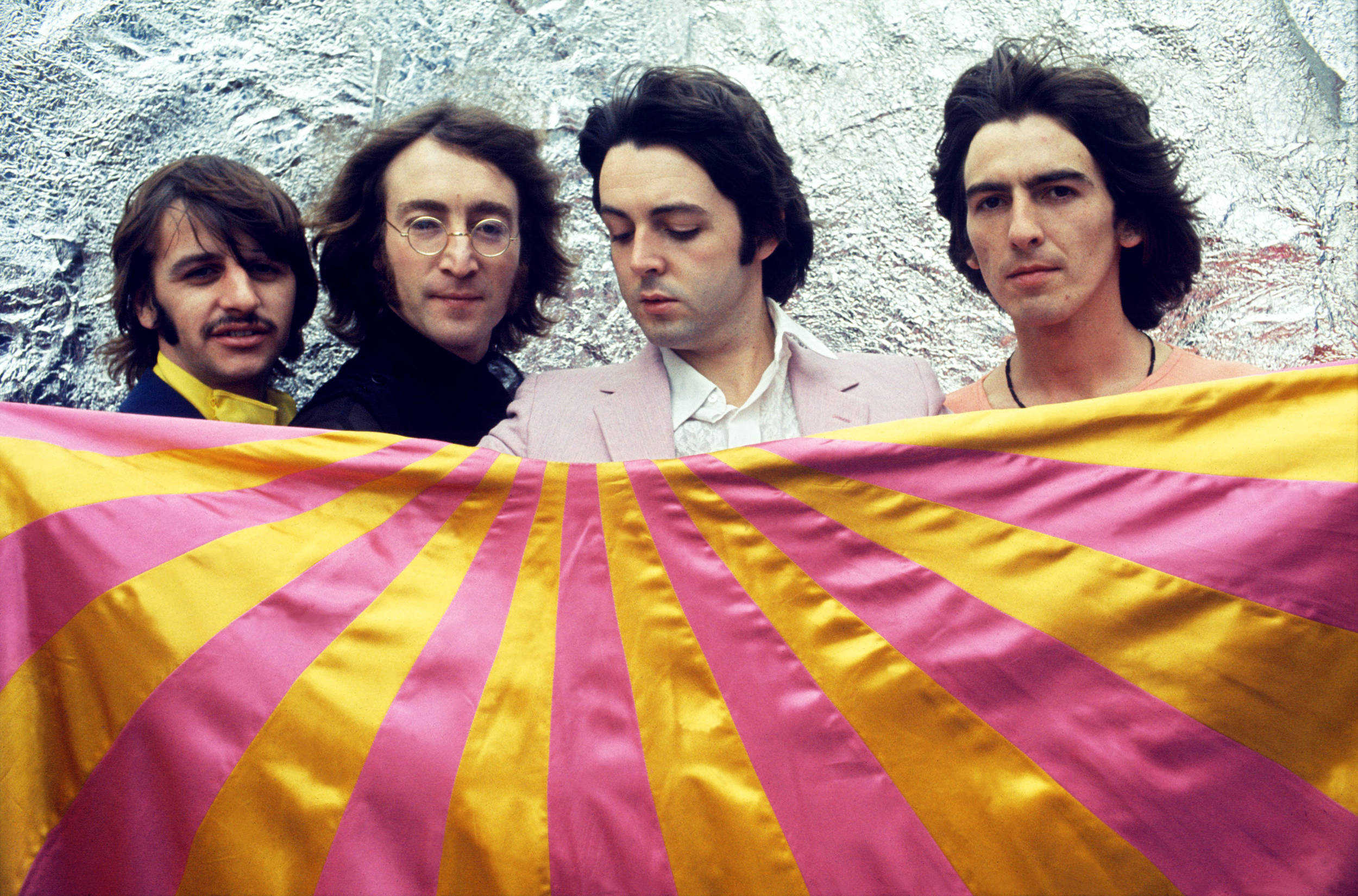 the beatles 桌布 壁紙 — 四名長髮男子在皺巴巴的銀色背景前舉著一面粉黃條紋旗幟。