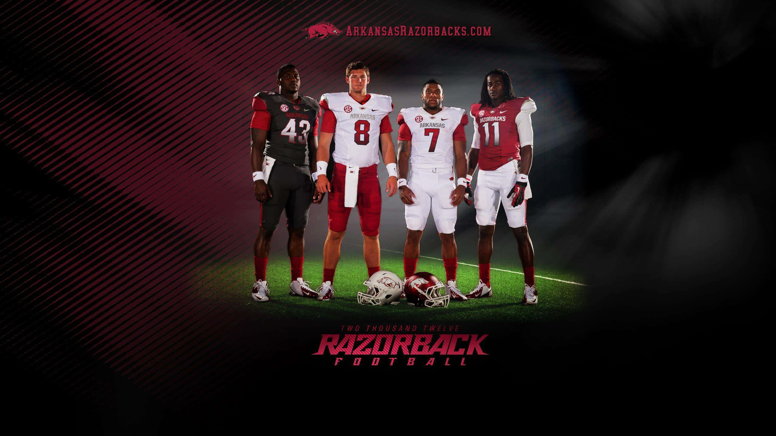 足球 桌布 壁紙 — 四名阿肯色剃刀鯨（Arkansas Razorbacks）美式足球員身穿紅白球衣，自信地站在聚光燈照射的球場上，頭盔放在腳邊。