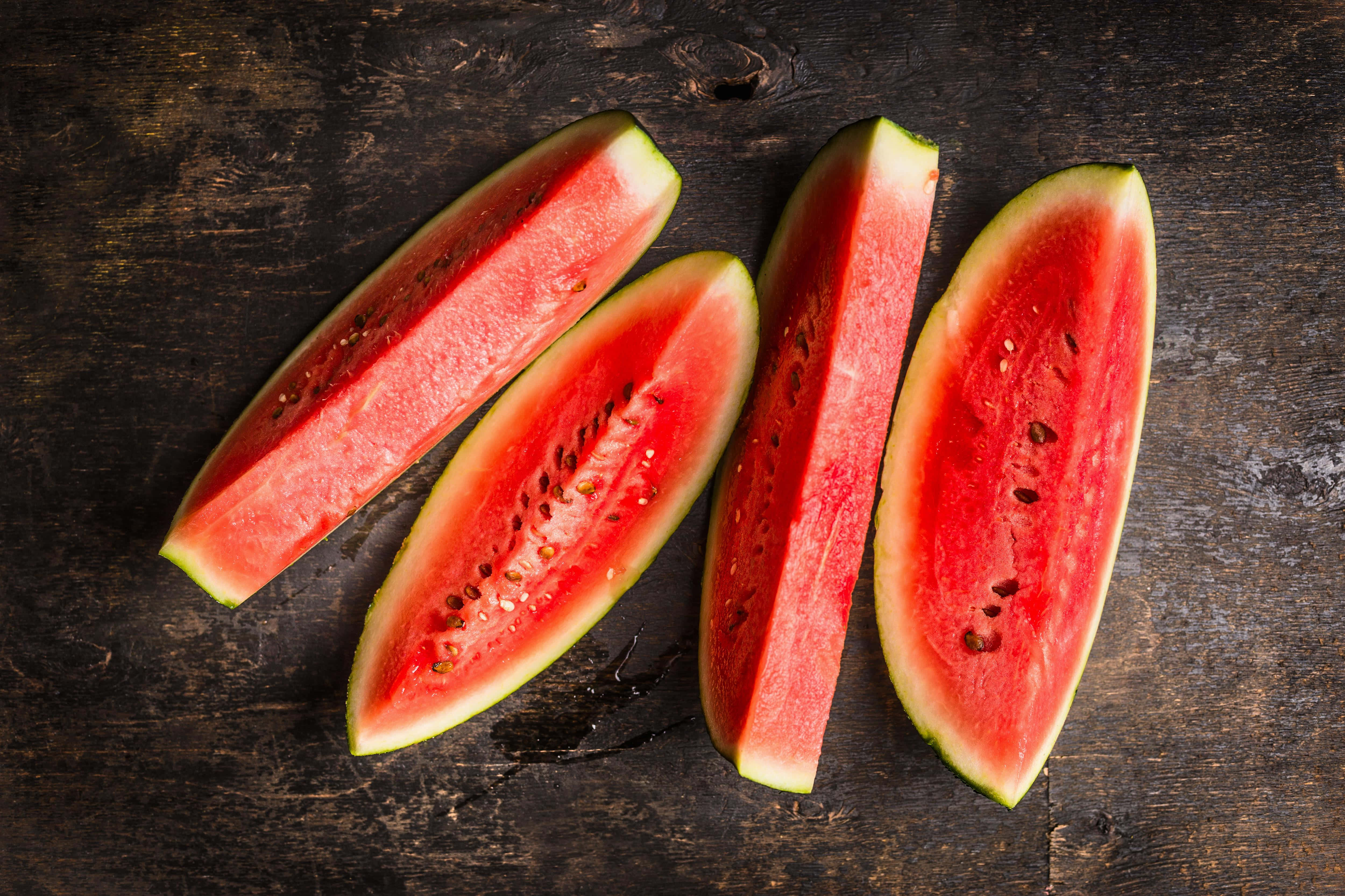 Watermelon 桌布 壁紙 — 四塊多汁的西瓜切片，帶有黑色種子，放在深色鄉村風木質表面上。