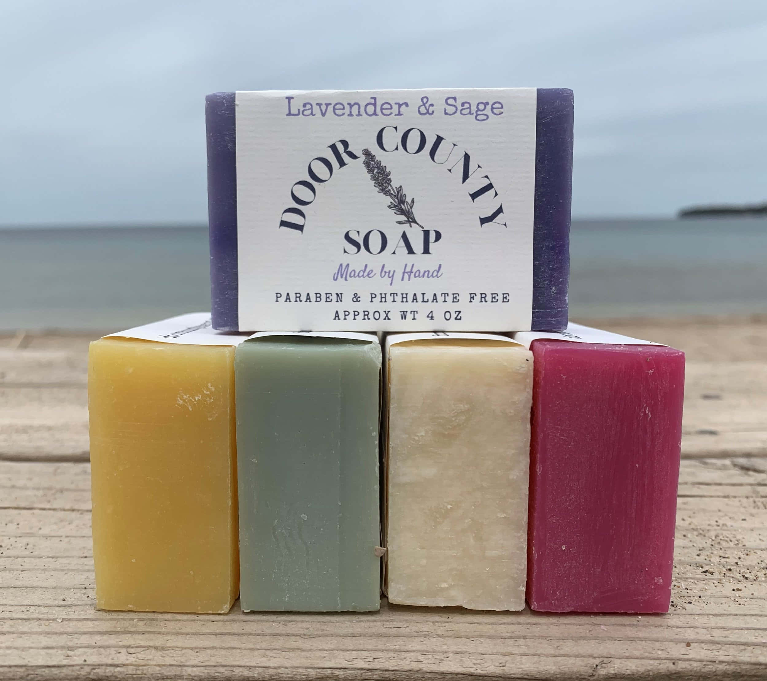 bath 桌布 壁紙 — 四塊色彩繽紛的手工皂，顏色有黃色、綠色、米色和洋紅色，放在經風化的木頭上，背景是柔和的海景。