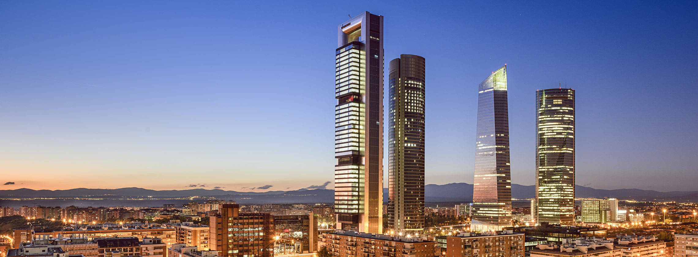 Madrid 桌布 壁紙 — 四座亮燈的摩天大樓在暮色天空中刺穿，下方是廣闊的城市景觀，遠山依稀可見。