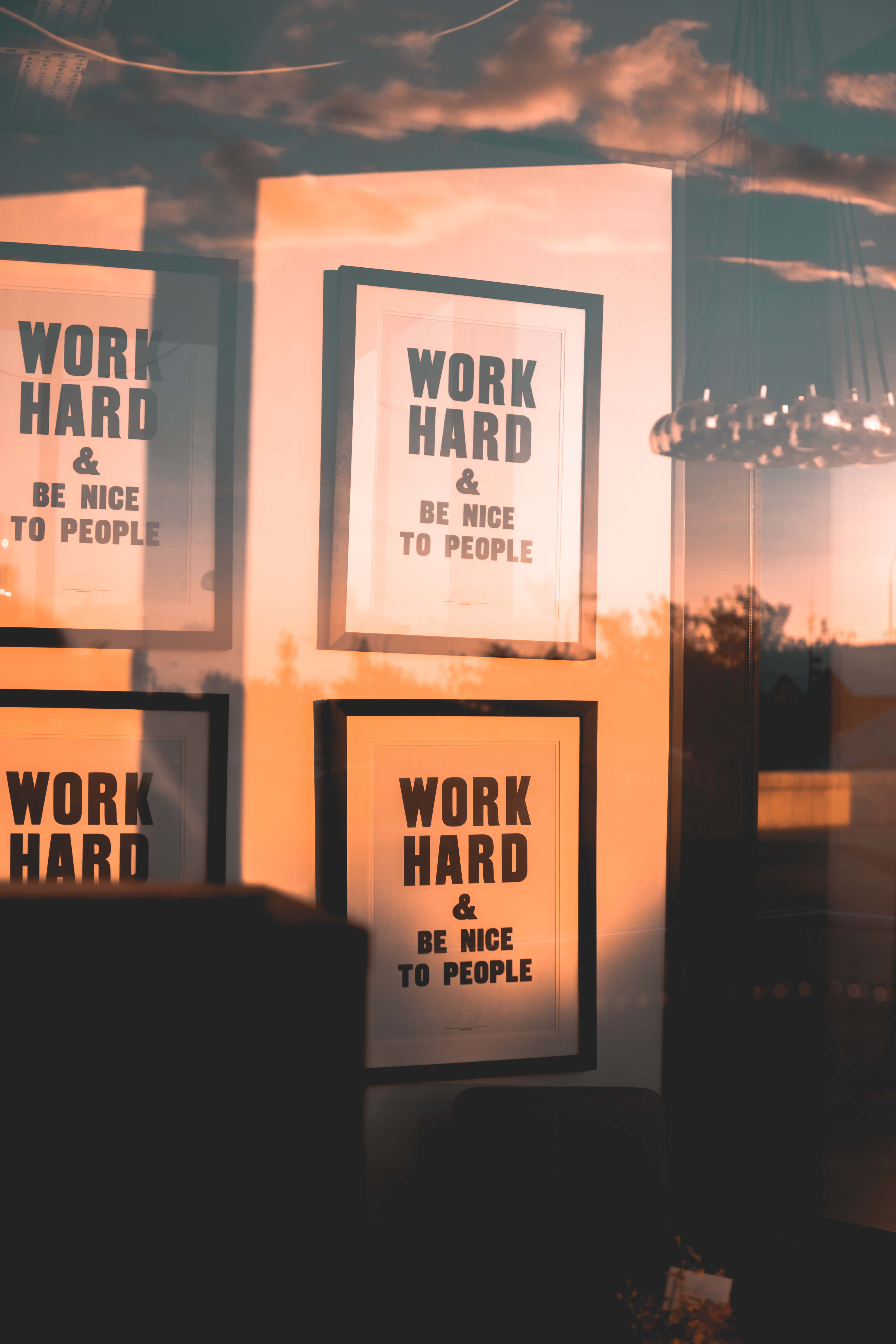 Hard Work 桌布 壁紙 — 四張裱框勵志海報，上面寫著「WORK HARD & BE NICE TO PEOPLE」，在玻璃反射的日落天空襯托下溫暖地發光。