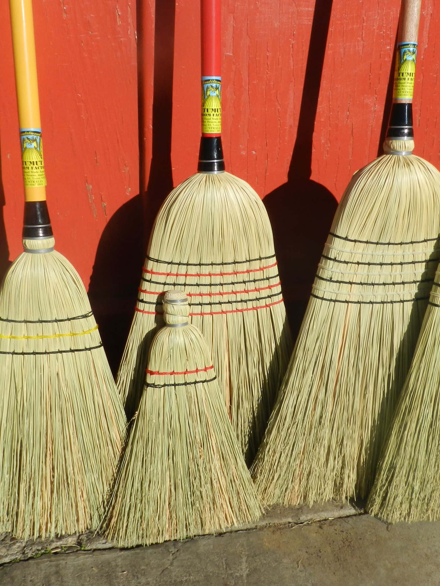 Broom 桌布 壁紙 — 四把彩色手柄的稻草掃帚靠在一面鮮紅色的木牆上，投下清晰的陰影。