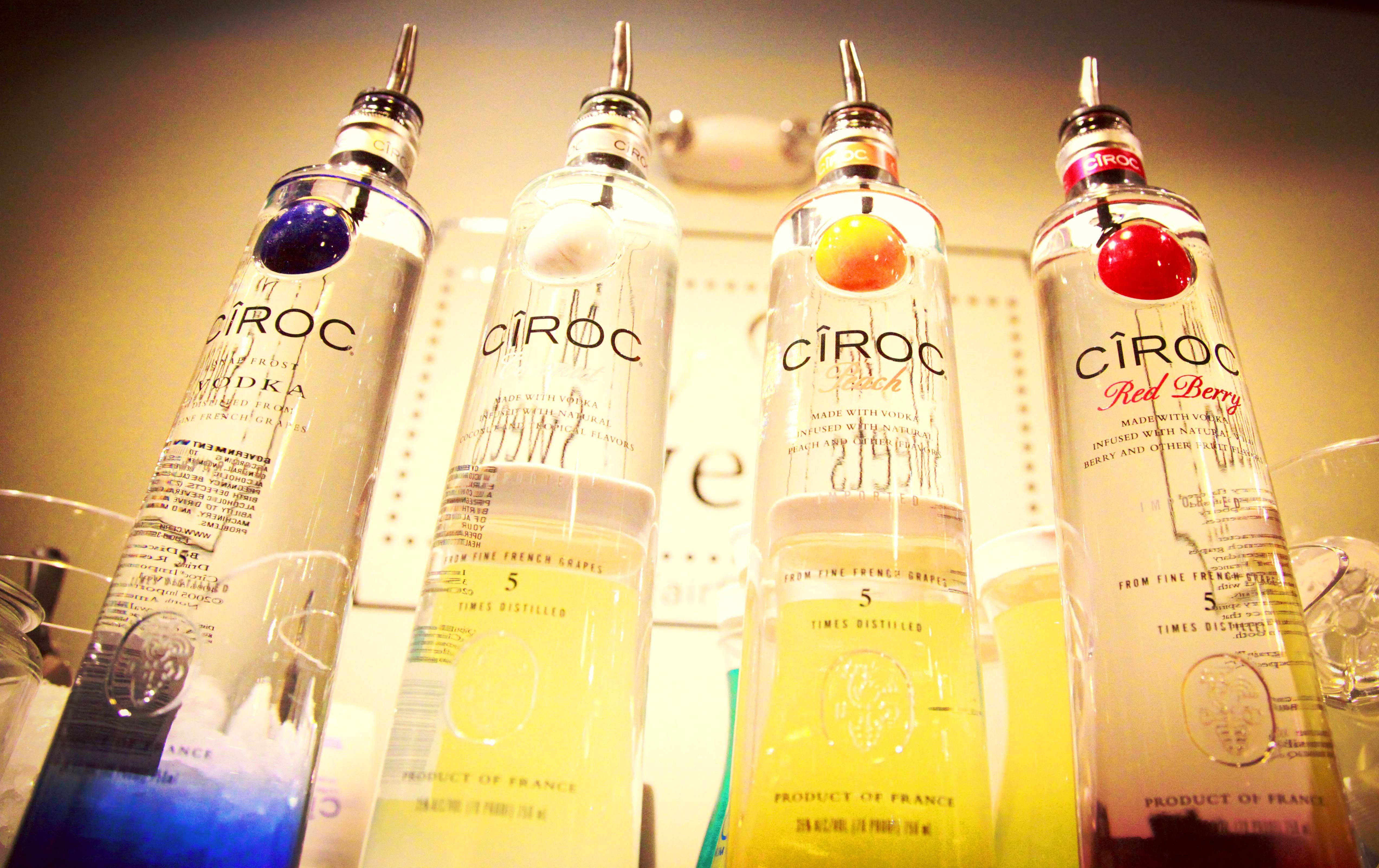 Drink 桌布 壁紙 — 四支時尚的Ciroc伏特加酒瓶配上水果裝飾，放置在溫暖燈光的酒吧環境中。