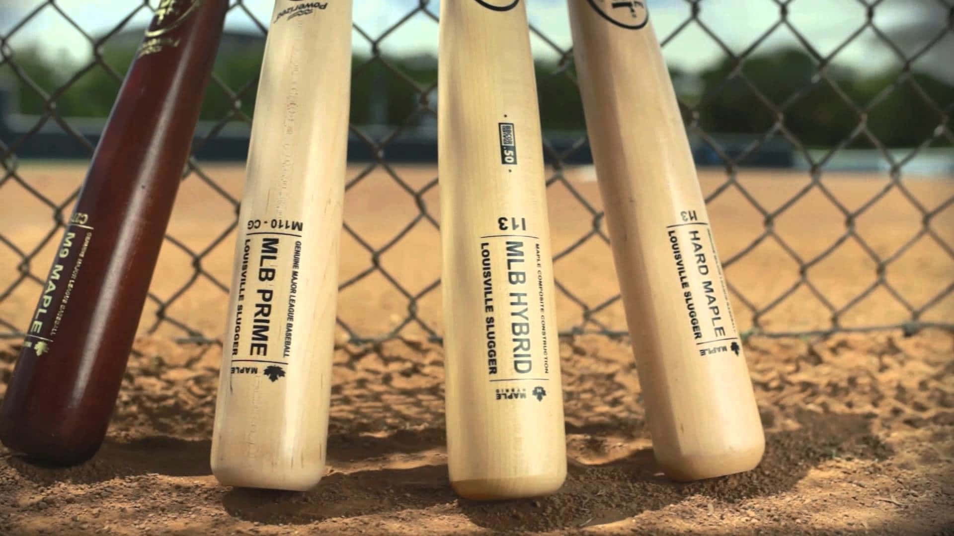 baseball bat 桌布 壁紙 — 四支棒球棍靠在陽光普照的泥地球場的鐵絲網圍欄上，背景是模糊的綠植。