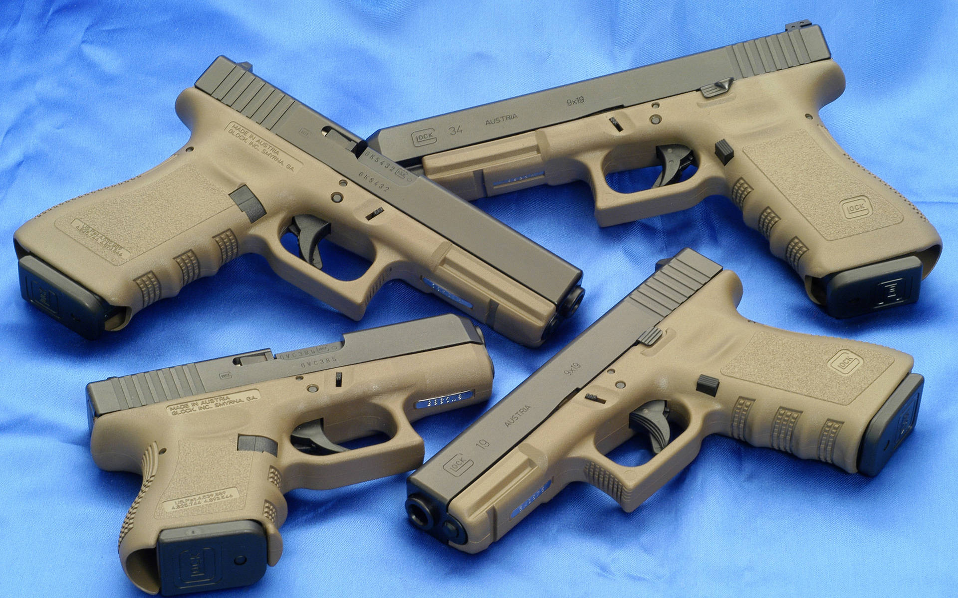 克拉克 桌布 壁紙 — 四支淺褐色 Glock 手槍排列在帶紋理的藍色布料背景上，呈現產品風格照片。