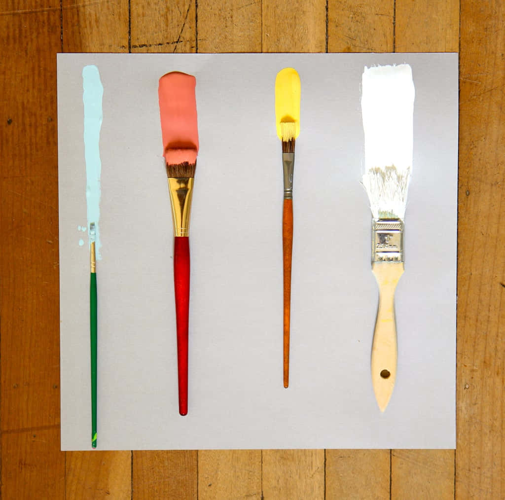 Paint Brush 桌布 壁紙 — 四支畫筆，刷毛上帶有淺藍色、珊瑚色、黃色和白色顏料，整齊地排列在木地板上的一張白色床單上。