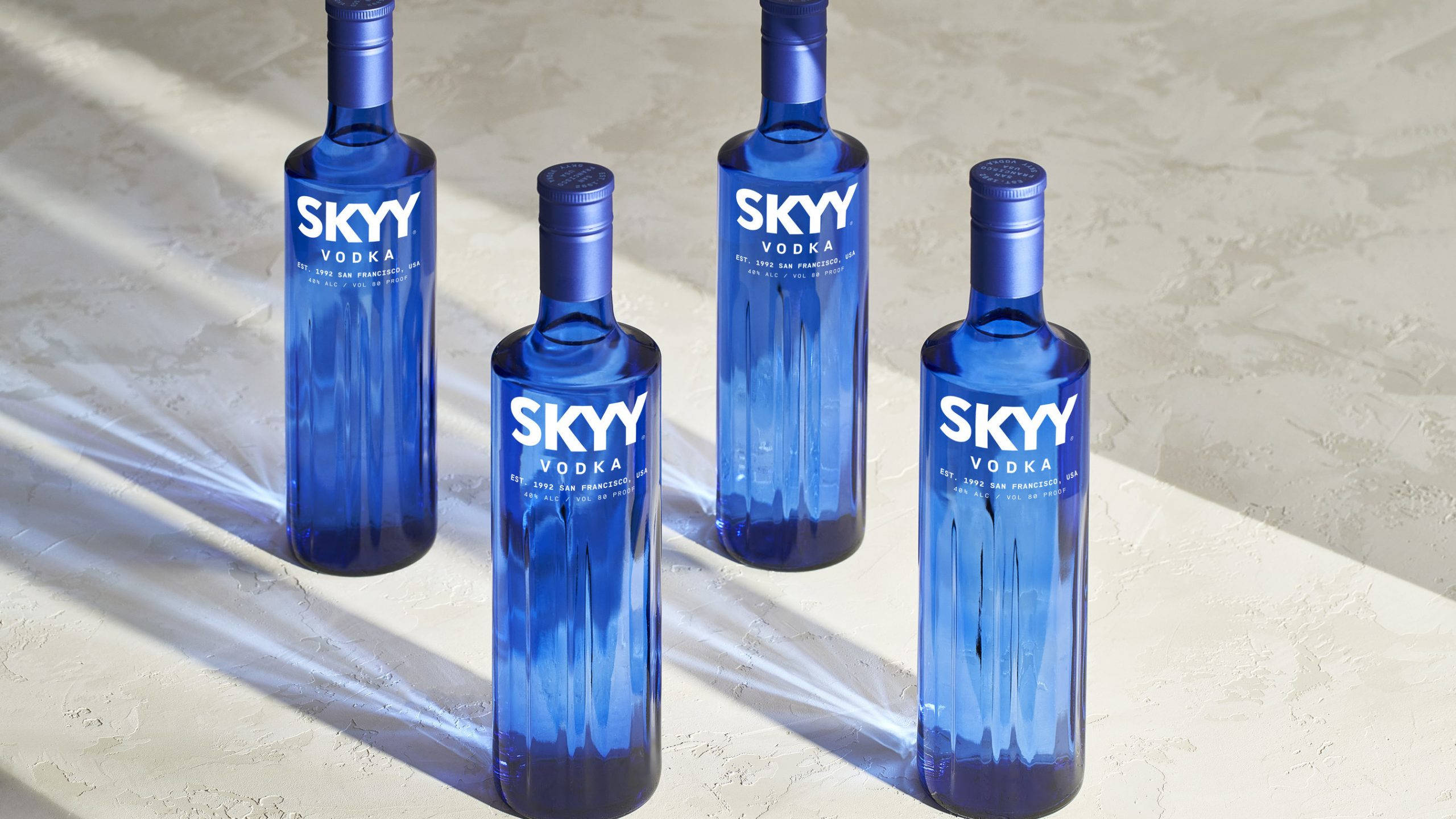 alcohol 桌布 壁紙 — 四支藍色SKYY伏特加酒瓶排列在紋理表面上，強烈的陽光投射出戲劇性的陰影。