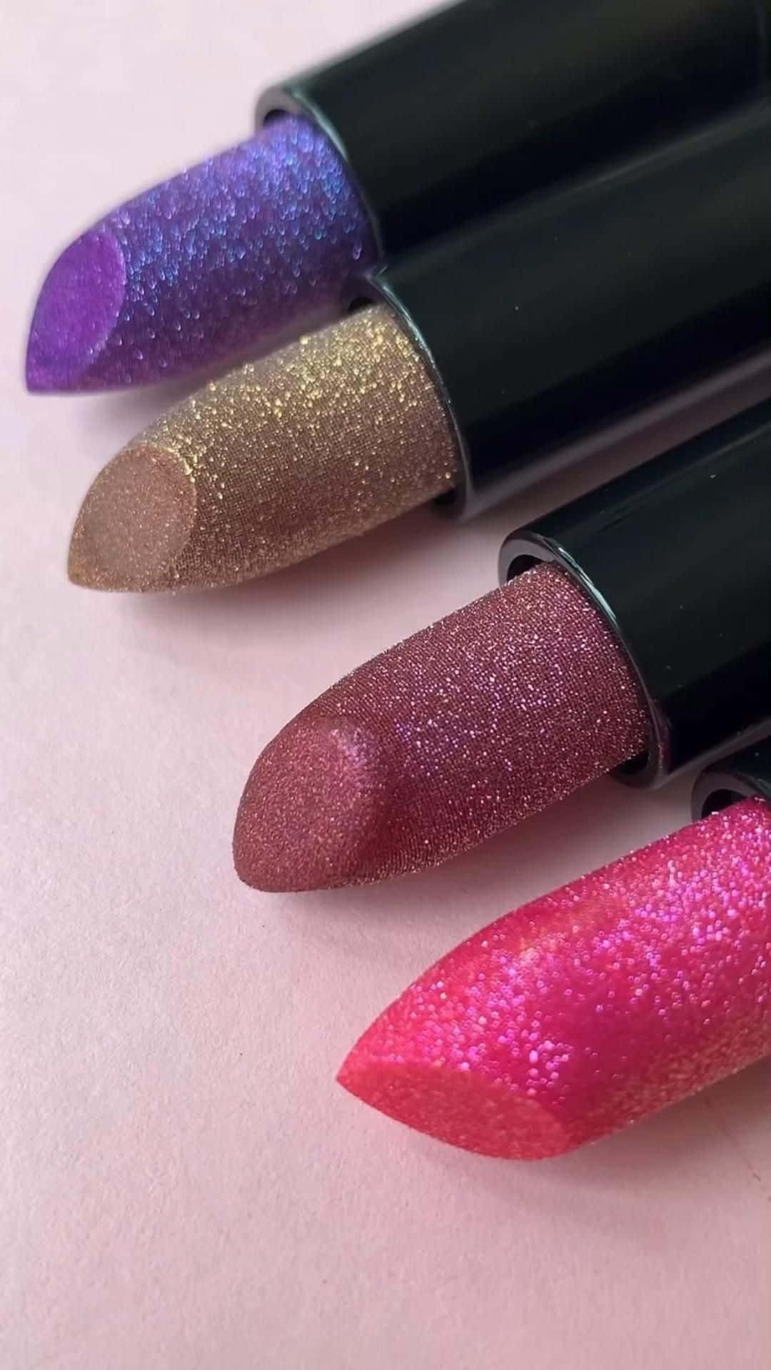 lipstick 桌布 壁紙 — 四支閃亮唇膏，分別為紫色、金色、玫瑰色和粉紅色調，在柔和的粉紅色背景上對角排列。