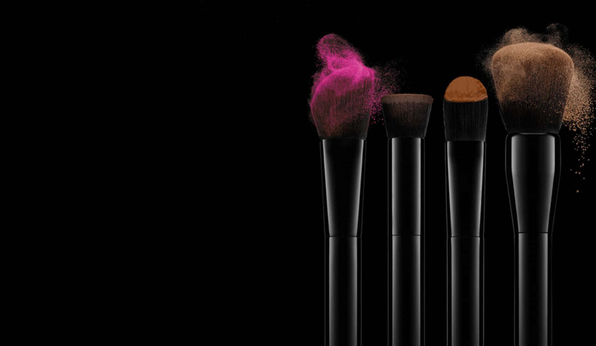 Cosmetics 桌布 壁紙 — 四支黑色化妝刷，刷毛上爆出粉紅色和棕色粉末，襯托在純黑背景上。