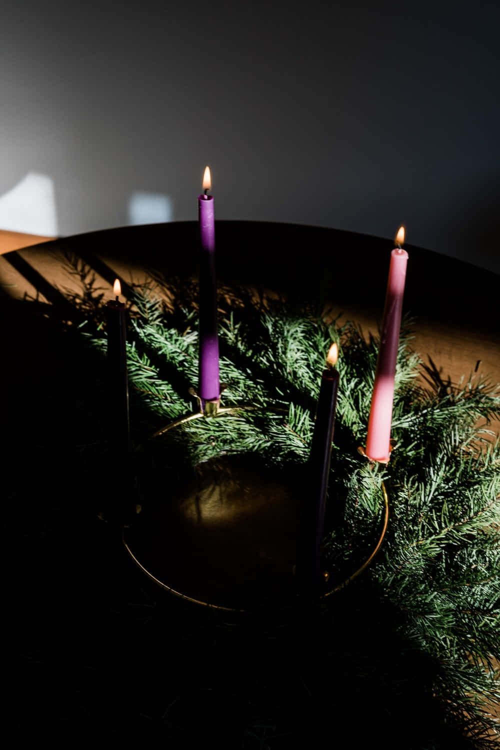 Advent 桌布 壁紙 — 四支點燃的蠟燭，呈現紫色、粉紅色和黑色色調，在一個圓形黃銅燭台上溫暖地發光，周圍環繞著常綠樹枝，光線昏暗。