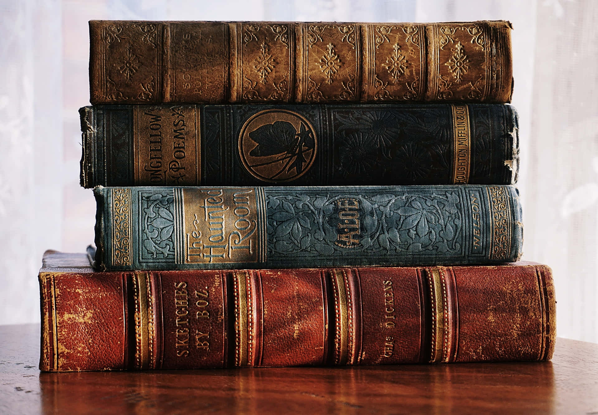 書本 桌布 壁紙 — 四本復古皮革裝訂書籍，帶有華麗的金色浮雕，堆放在拋光木桌上，靠近薄紗窗簾。