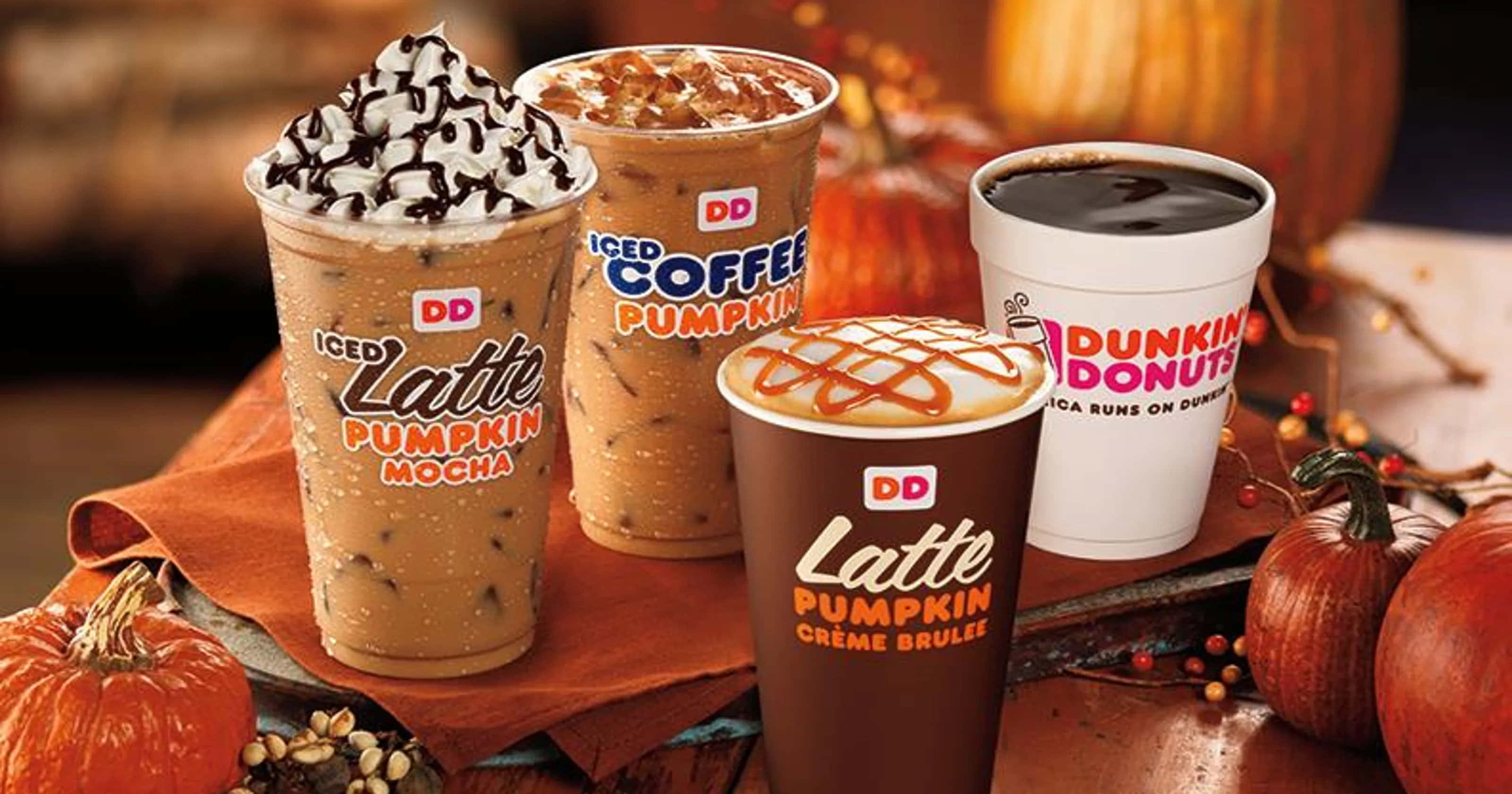 Dunkin Donuts 桌布 壁紙 — 四杯Dunkin' Donuts南瓜風味飲品，上面有鮮奶油、焦糖和巧克力醬，周圍有南瓜和秋季裝飾。
