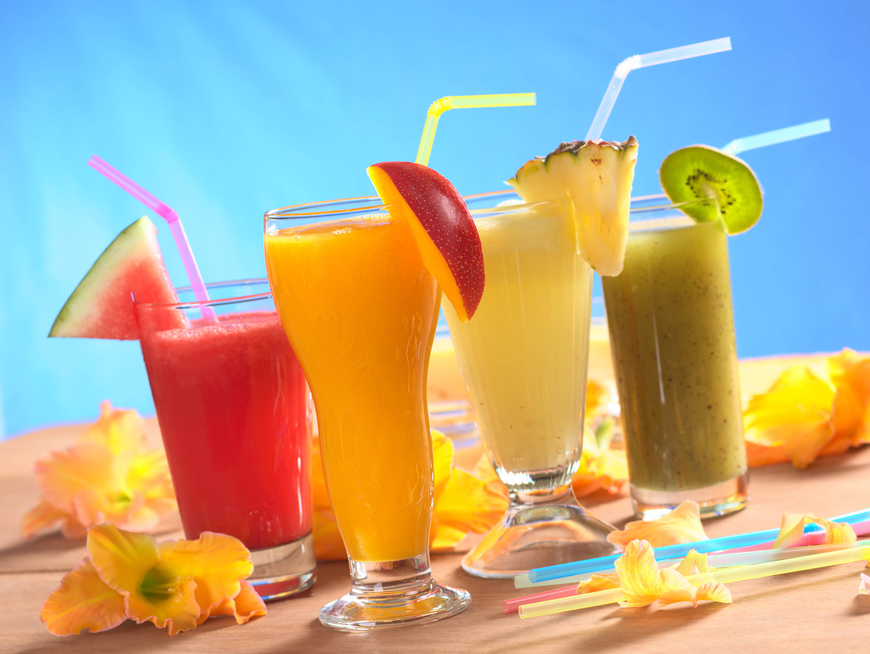 Smoothie 桌布 壁紙 — 四杯充滿活力的熱帶冰沙，裝在高腳杯中，飾以水果片和彩色吸管，背景為明亮的藍色。