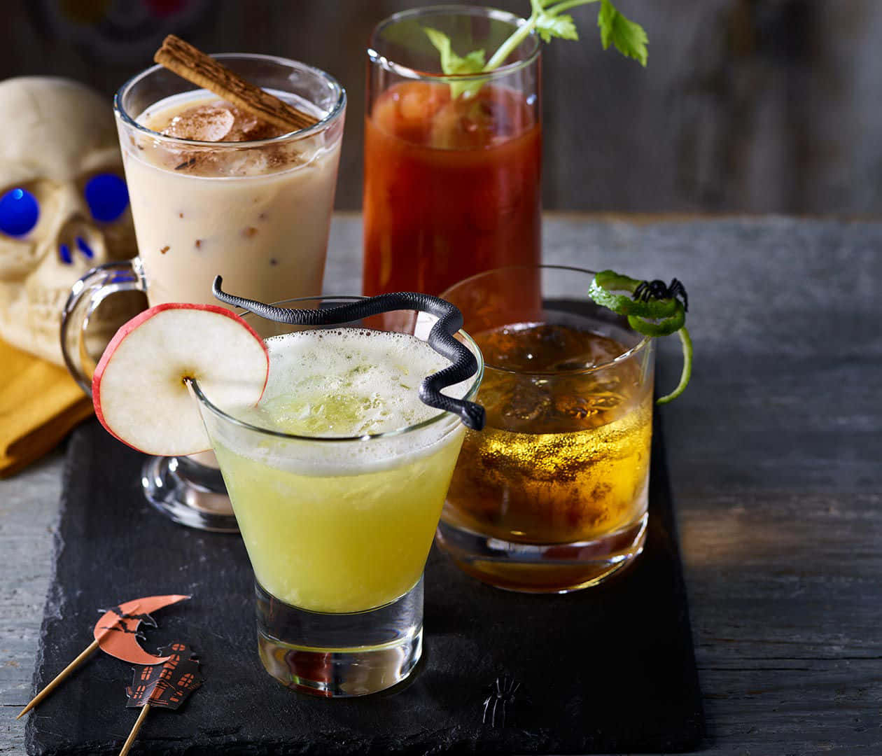Halloween Drinks 桌布 壁紙 — 四杯萬聖節恐怖雞尾酒，配有蘋果片和假蜘蛛等裝飾，放在一個深色石板托盤上，旁邊有一個骷髏頭。