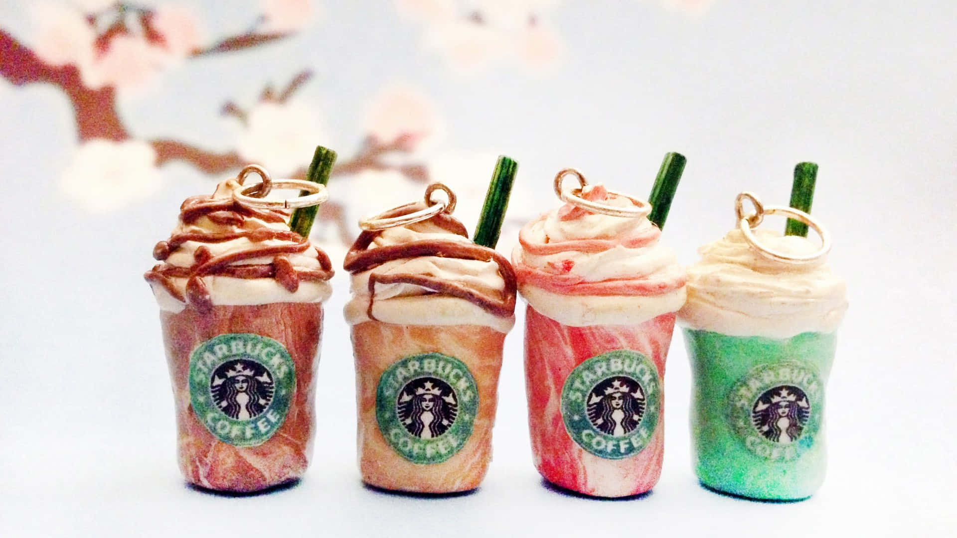 Aesthetic Starbucks 桌布 壁紙 — 四杯迷你星巴克飲品，配有鮮奶油和吸管，排成一排，背景是柔和模糊的櫻花。