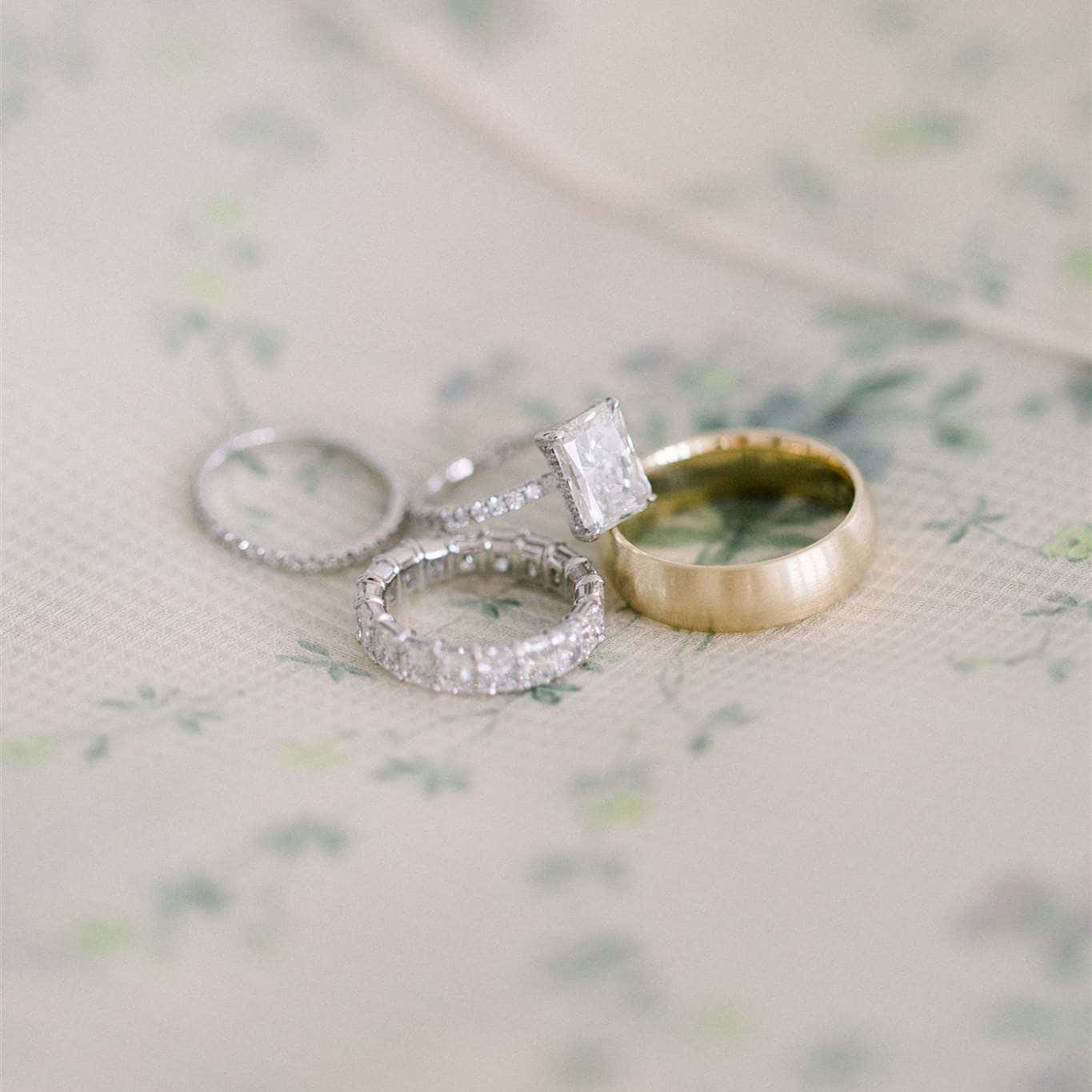 Wedding Rings 桌布 壁紙 — 四枚優雅的結婚戒指，包括一枚金戒指和鑲鑽戒指，放在柔軟的花卉布料上。