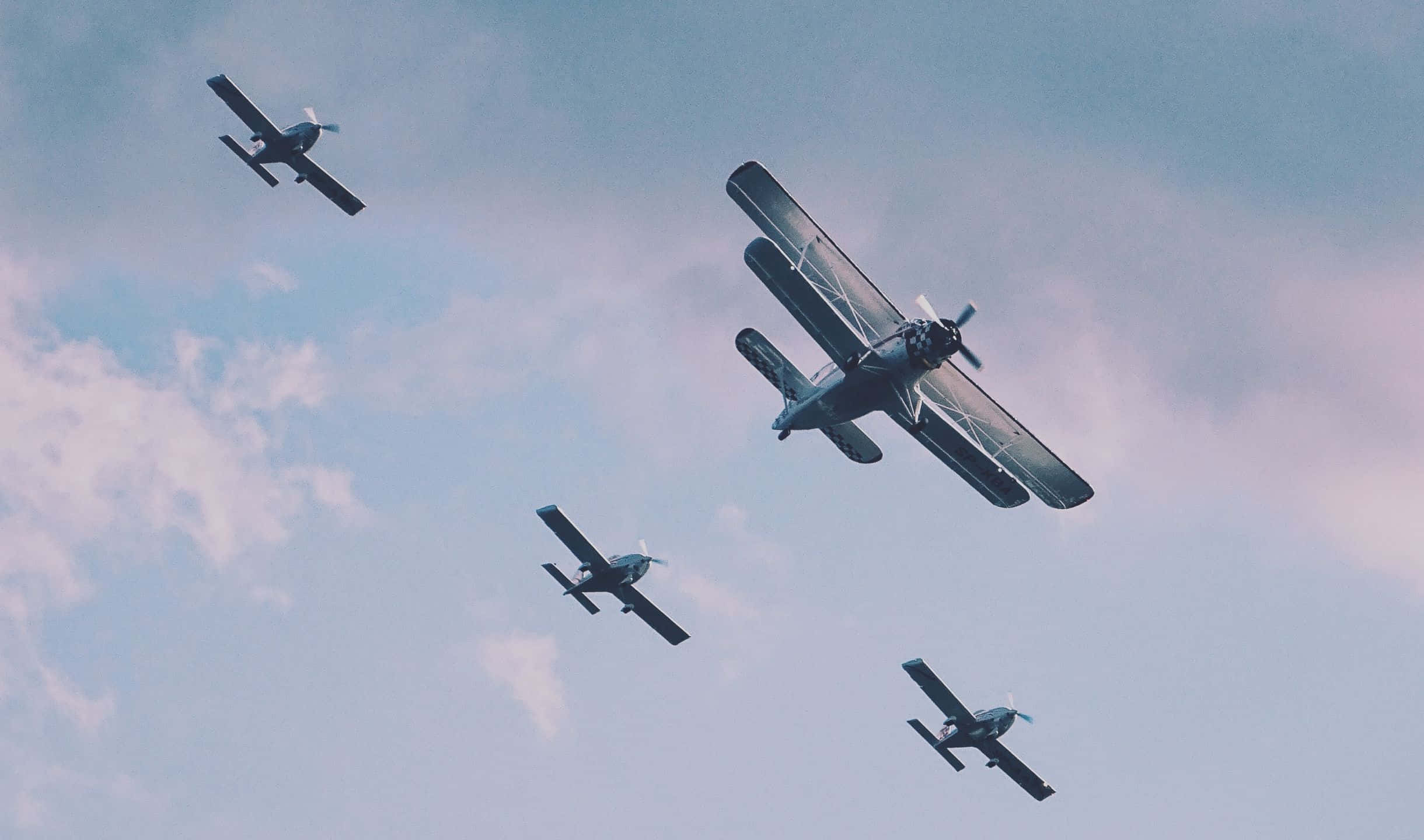 2440x1440 Small Planes 桌布 壁紙 — 四架復古雙翼機在柔和粉彩天空及縷縷白雲背景下編隊翱翔。