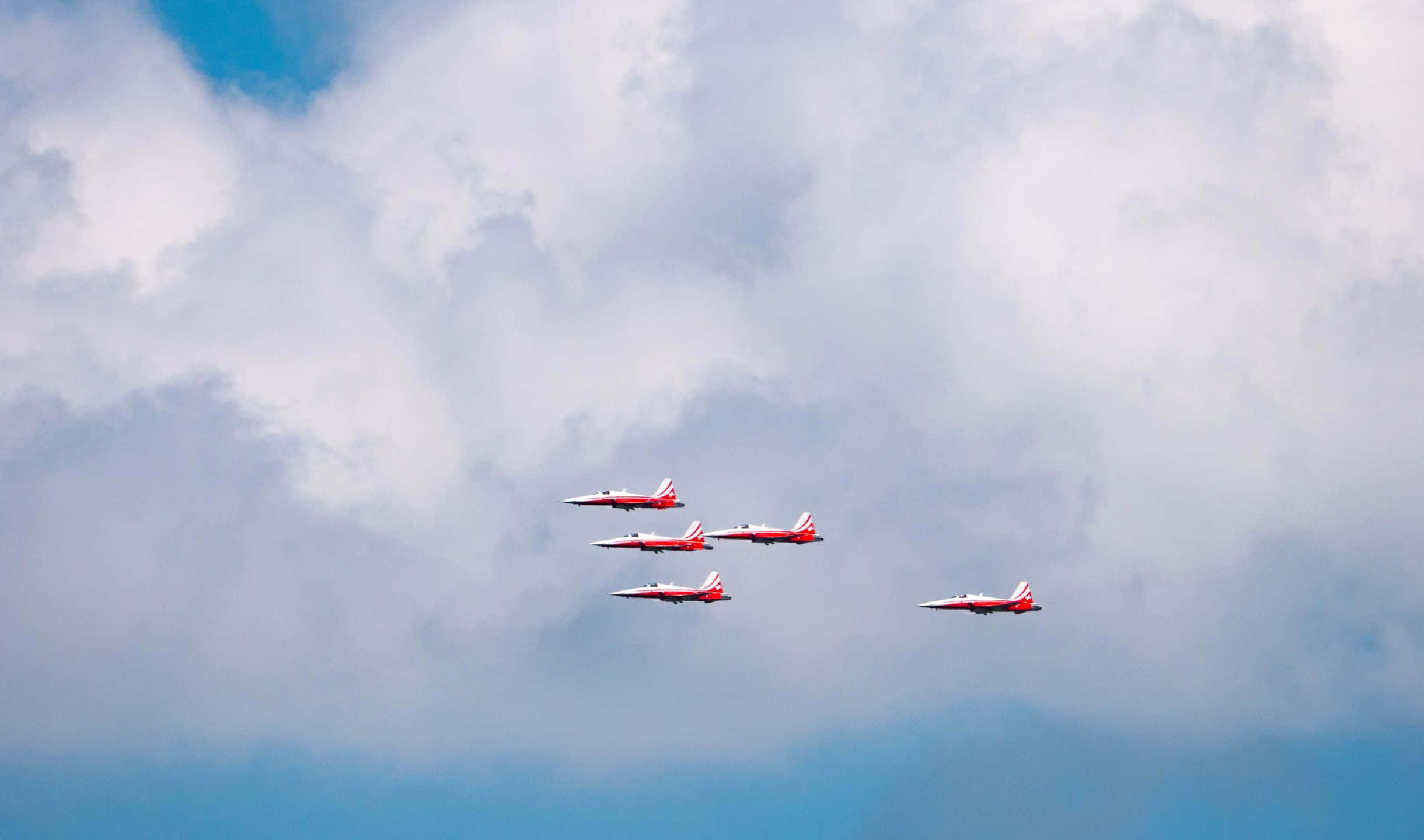 2440x1440 Small Planes 桌布 壁紙 — 四架紅白相間的噴射機以緊密編隊飛行，穿過充滿蓬鬆白雲和藍色斑塊的戲劇性天空。