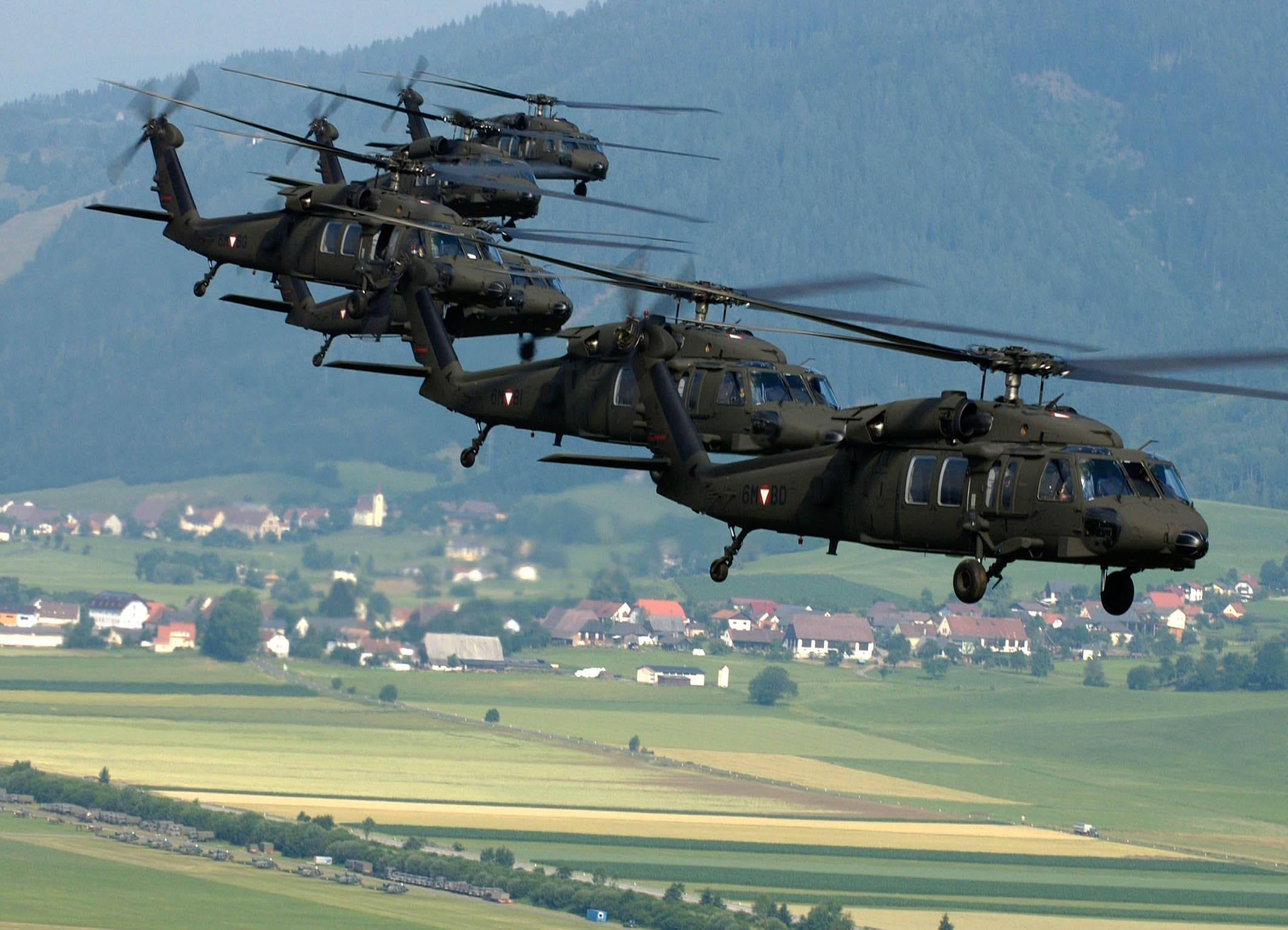 Black Hawk Helicopter 桌布 壁紙 — 四架軍用直升機編隊飛越綠色山谷，下方有村莊，遠處有森林覆蓋的山丘。