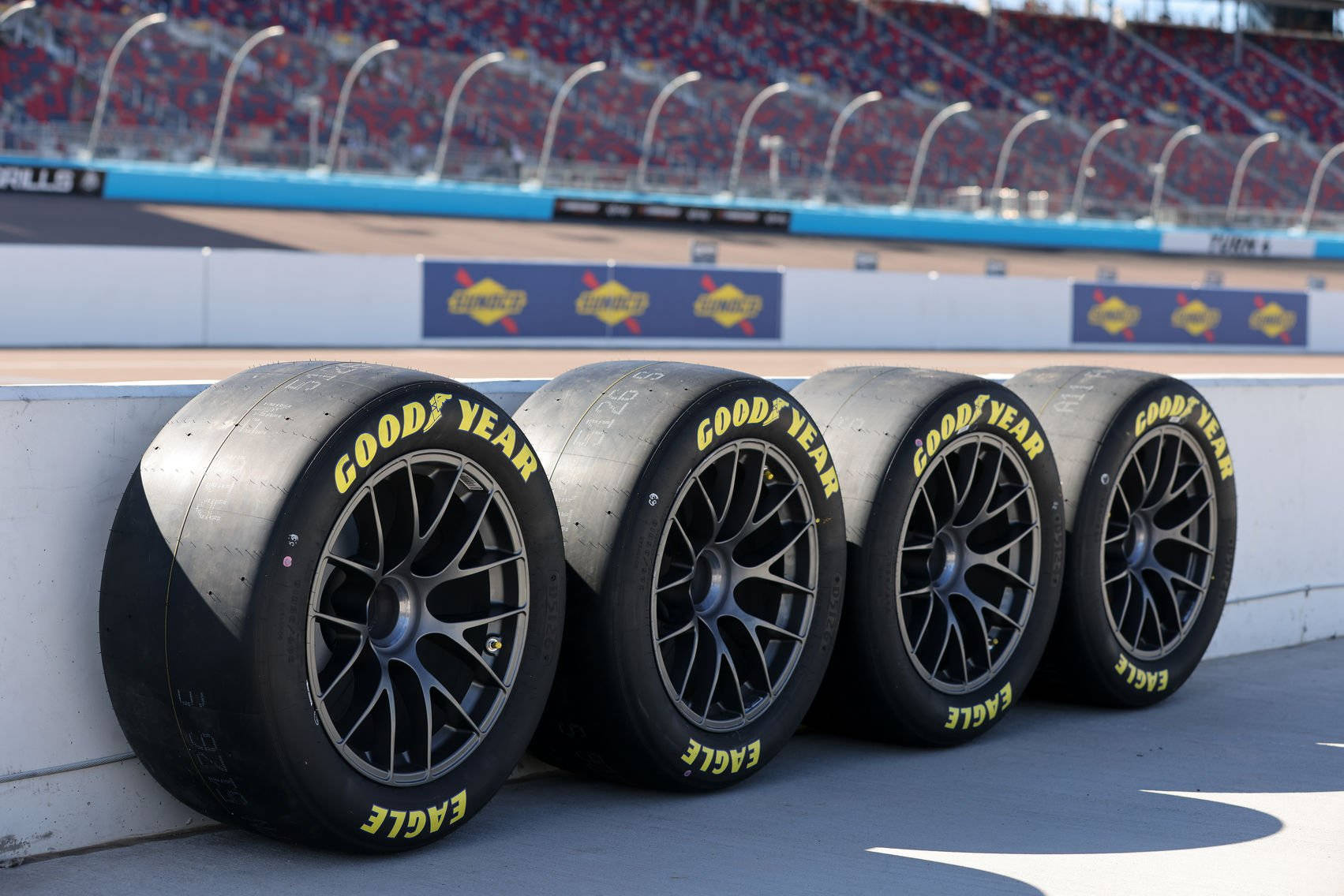 Racing 桌布 壁紙 — 四條Goodyear Eagle賽車輪胎靠在陽光明媚的賽車場的混凝土護欄上，背景是空蕩蕩的看台。