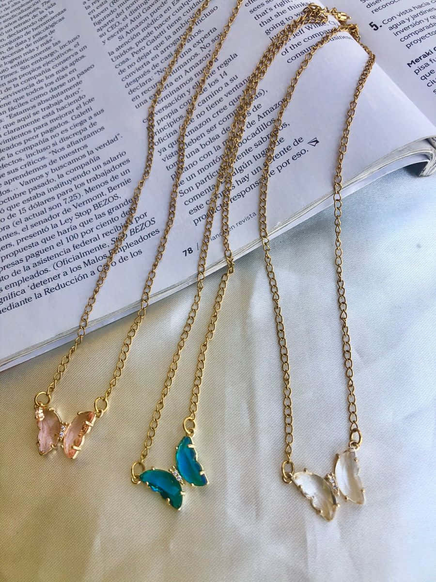 Butterfly Jewelry 桌布 壁紙 — 四條帶有粉紅、蜜桃、青綠和白色蝴蝶吊墜的金鏈優雅地放在一本打開的書本和白色布料上。