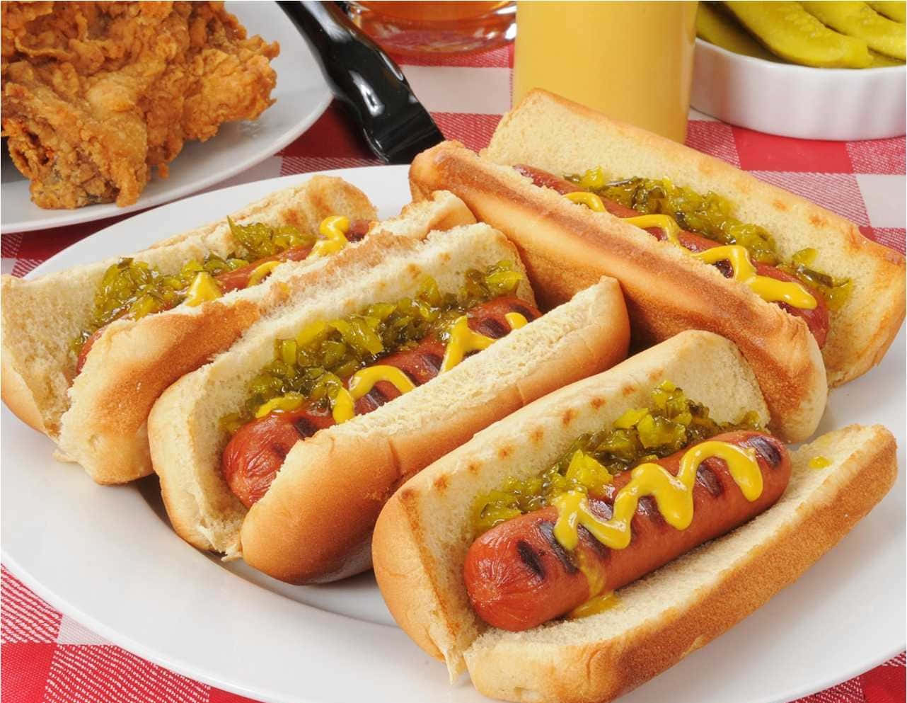 Hot Dog 桌布 壁紙 — 四條烤熱狗麵包配上芥末和調味醬，放在白色盤子上，旁邊有炸雞和酸黃瓜，置於紅色格子桌布上。