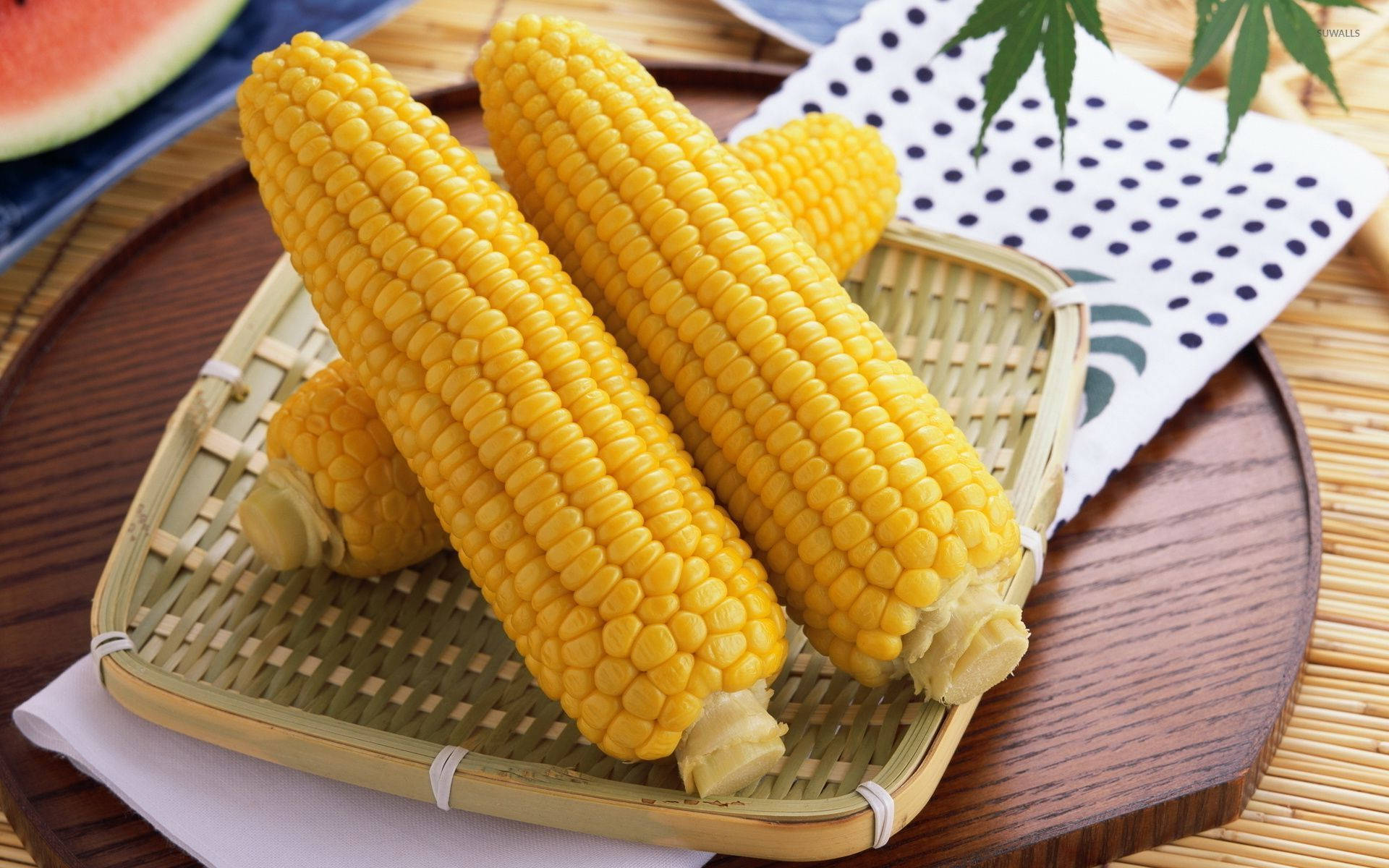 Food Photography 桌布 壁紙 — 四條鮮黃色的玉米棒放在一個編織竹籃裡的木托盤上，旁邊有一塊西瓜片和一片綠葉。