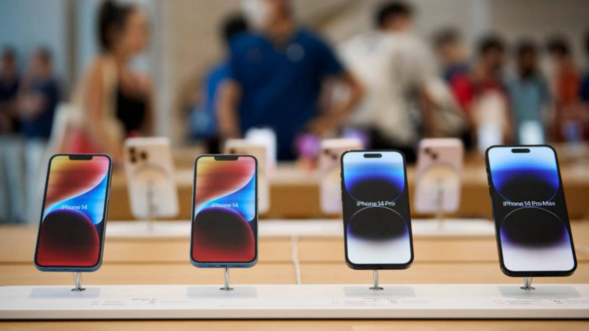 apple iPhone default 桌布 壁紙 — 四款 iPhone 14 型號直立展示在零售店的支架上，背景有模糊的顧客。