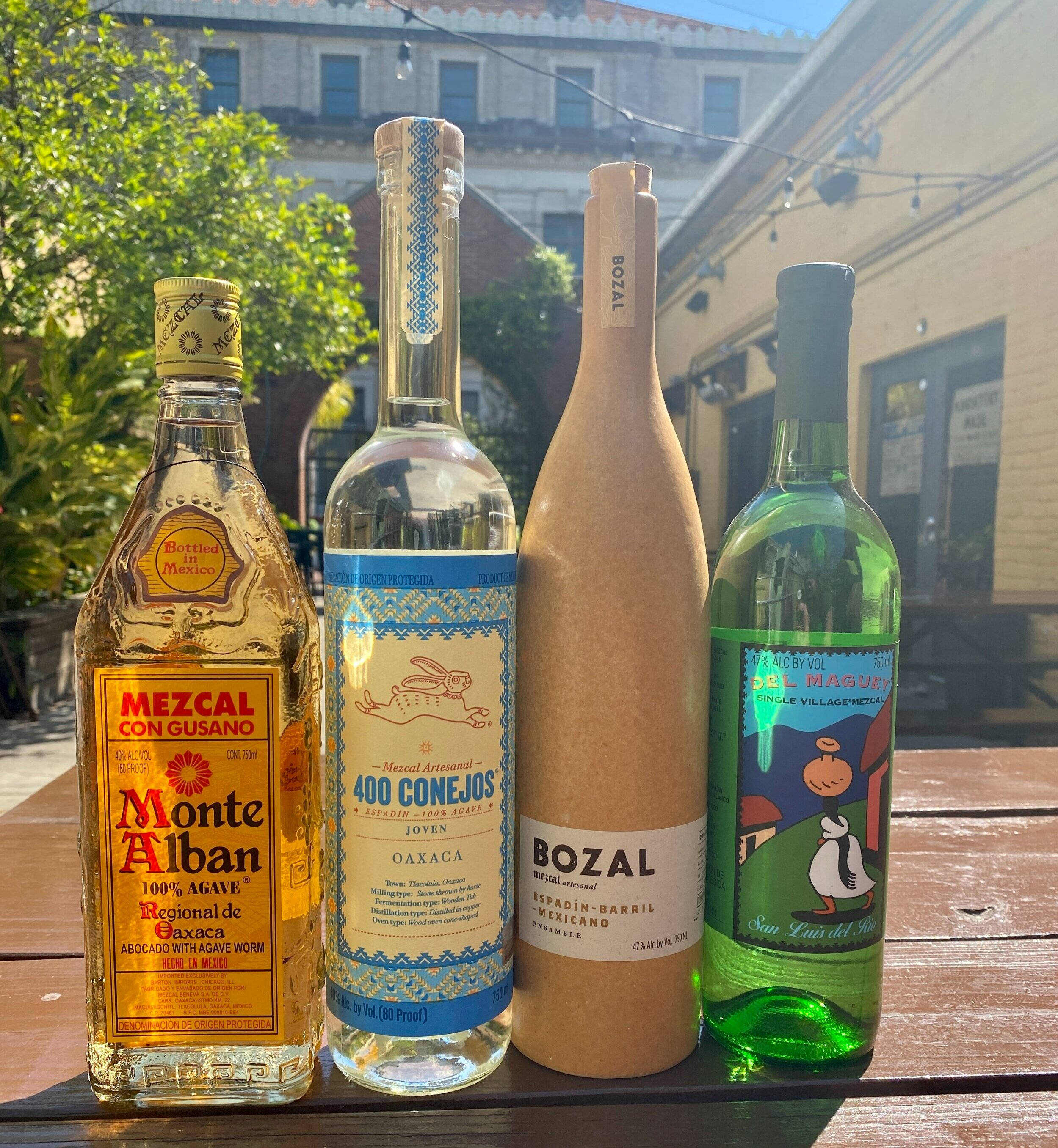 Drink 桌布 壁紙 — 四款不同的墨西哥烈酒瓶放在木桌上，背景是陽光普照的戶外庭院，有建築物和綠植。