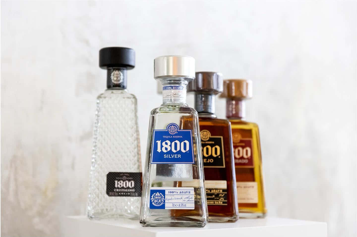 alcohol 桌布 壁紙 — 四款不同顏色和設計的1800龍舌蘭酒瓶整齊地排列在白色表面上，背景是柔軟的紋理牆壁。