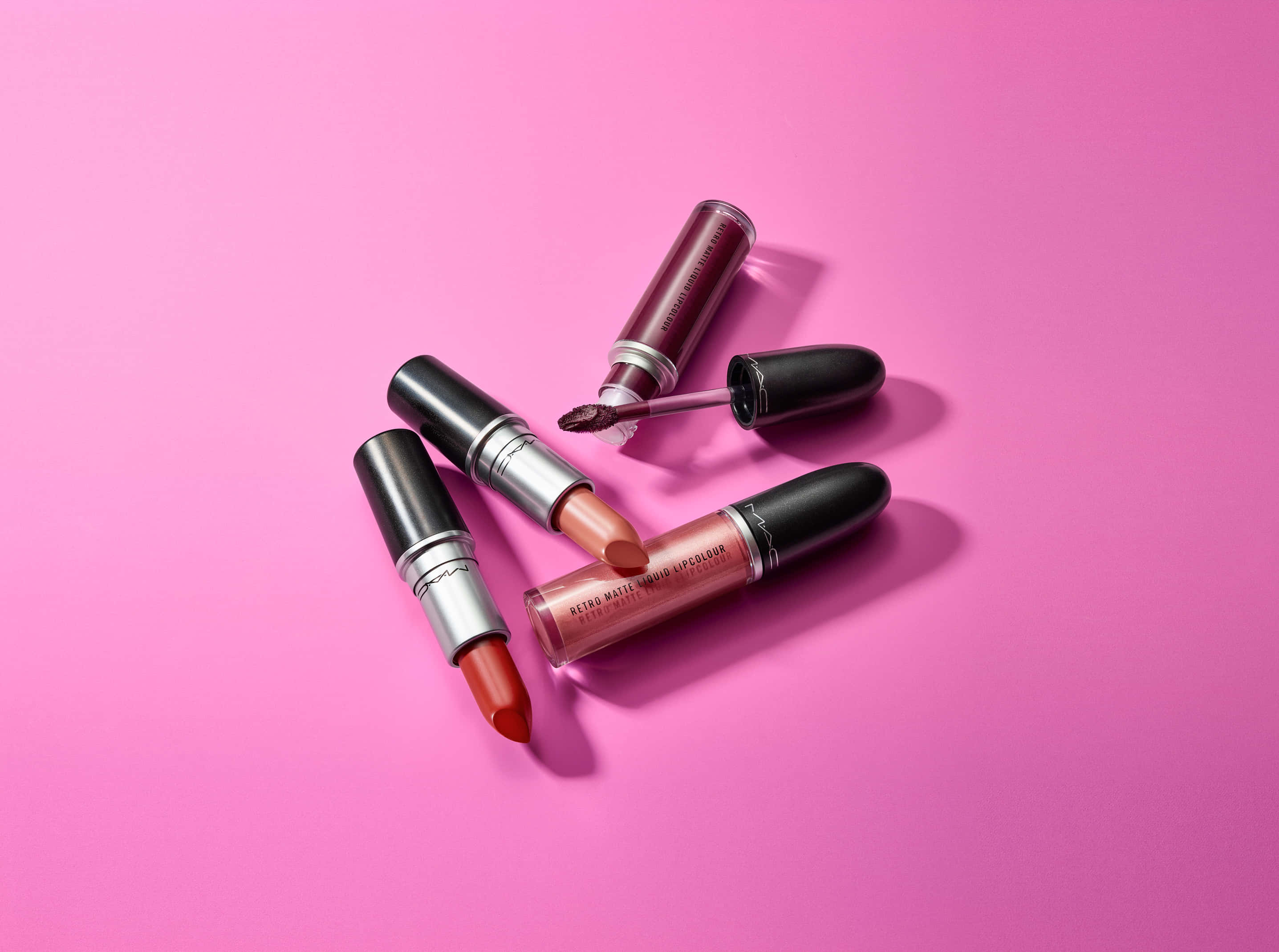 Mac Cosmetics 桌布 壁紙 — 四款暖色調的MAC唇部產品散落在充滿活力的粉紅色背景上，帶有柔和陰影。